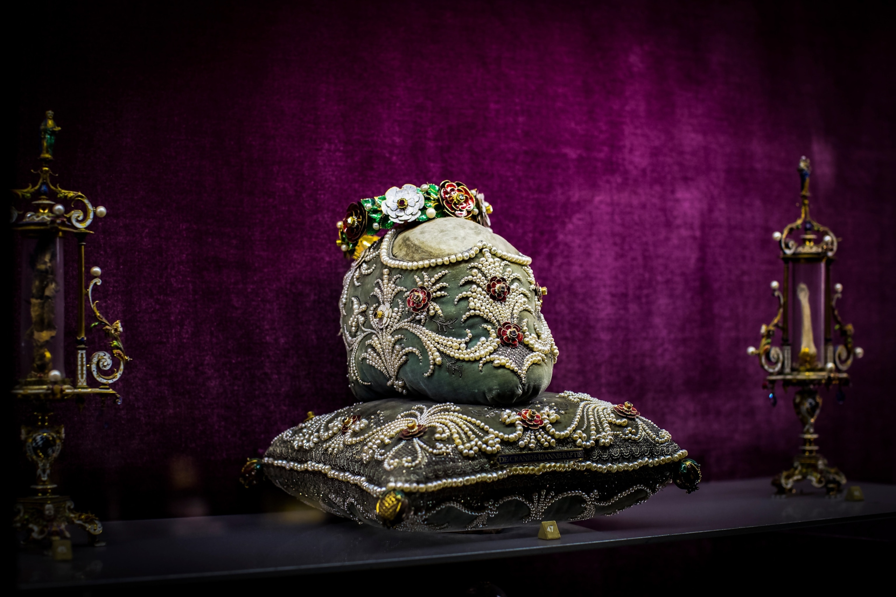 A skull-shaped object wrapped in mossy-green velvet that is decorated with pearls and flower ornaments sitting atop an equally ornamental velvet pillow.