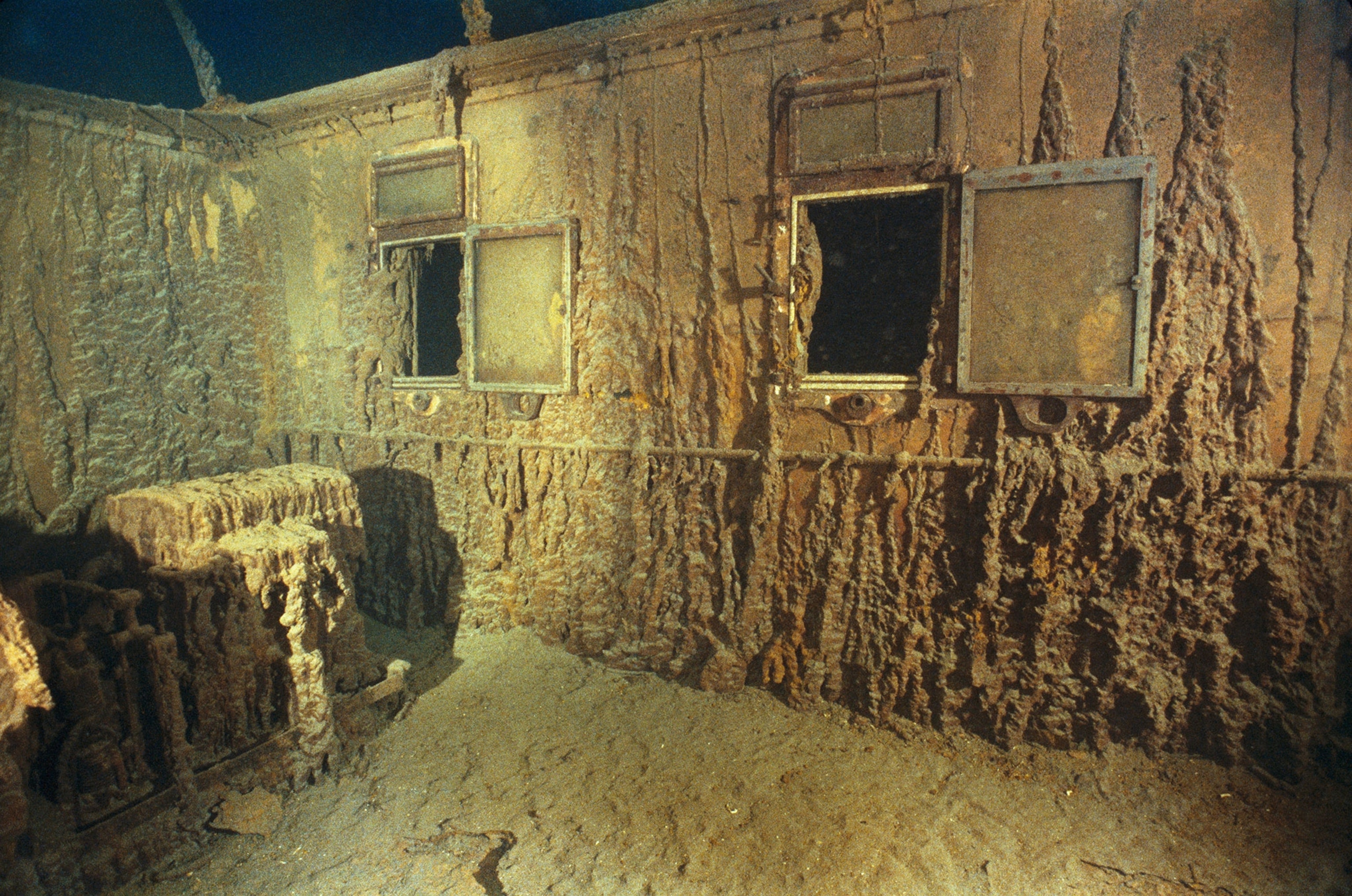 The inside of a room on the titanic shown covered in elements from the ocean. The wall shown has two open windows.