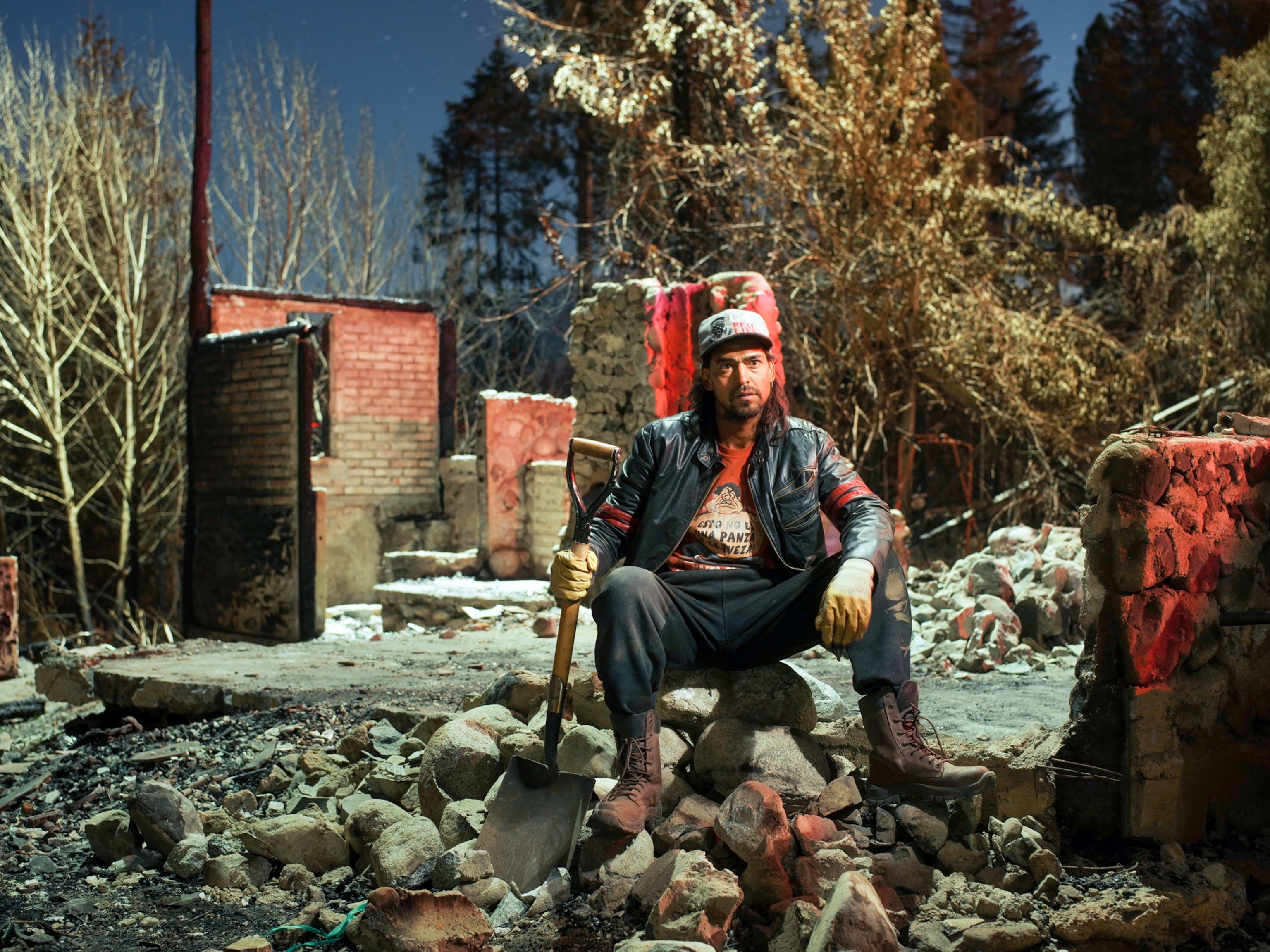 a man sits on the rubble of his burnt home holding a shovel