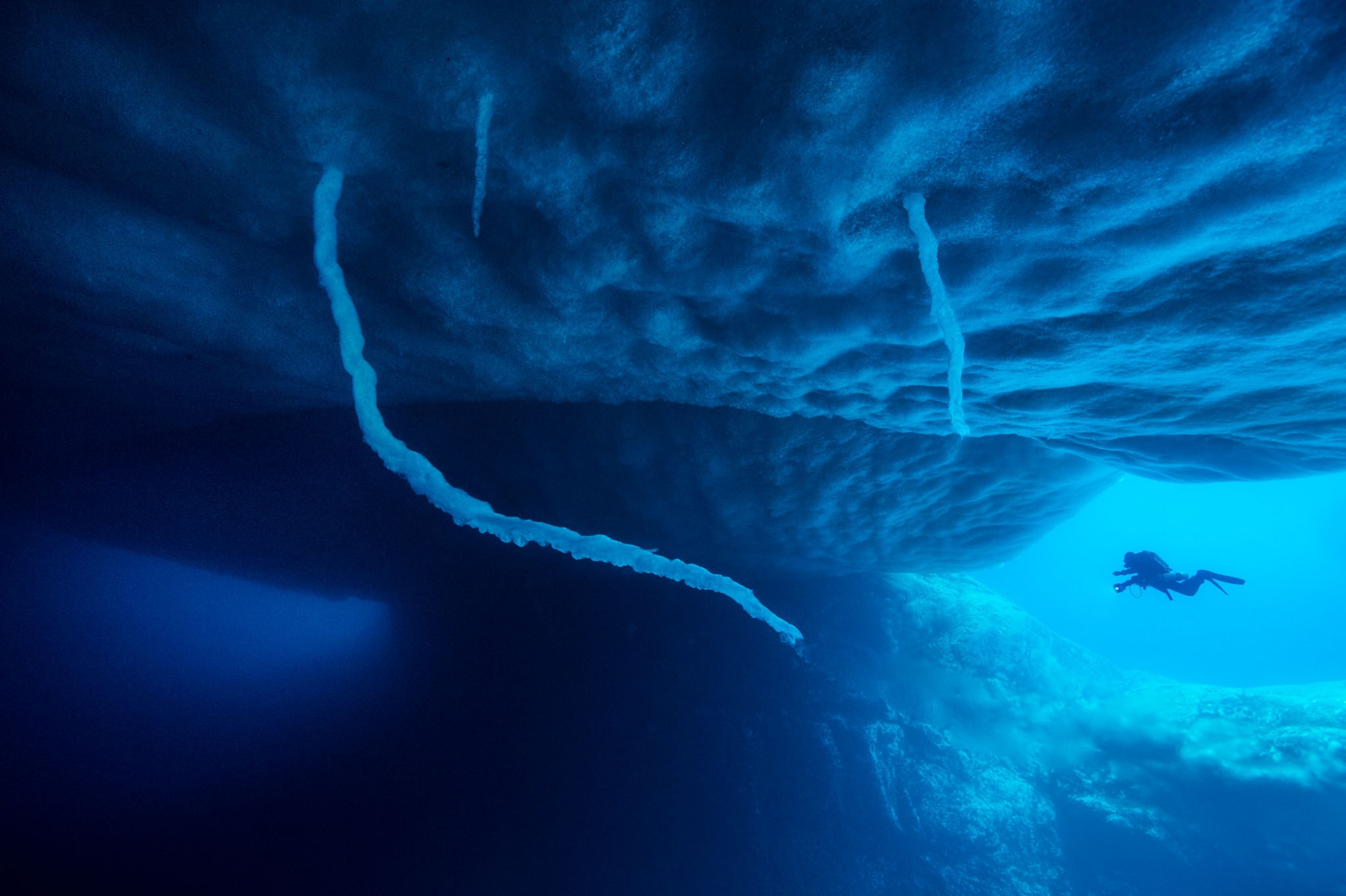 a lone diver swimming under sea ice in rich blue water