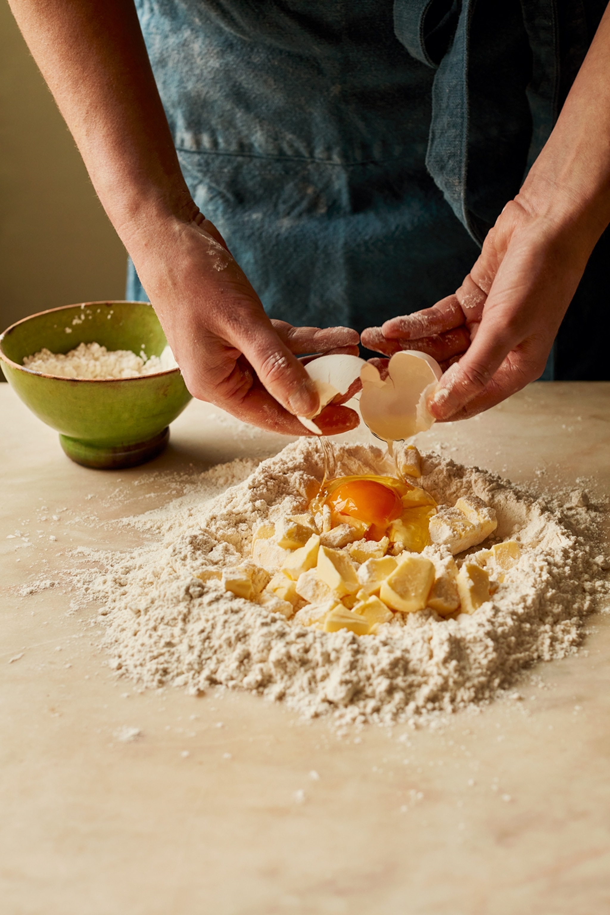 A pile of flour on a countertop with cubed butter in the middle. A man is cracking an egg onto the butter and flour mixture, whilst wearing a blue apron.