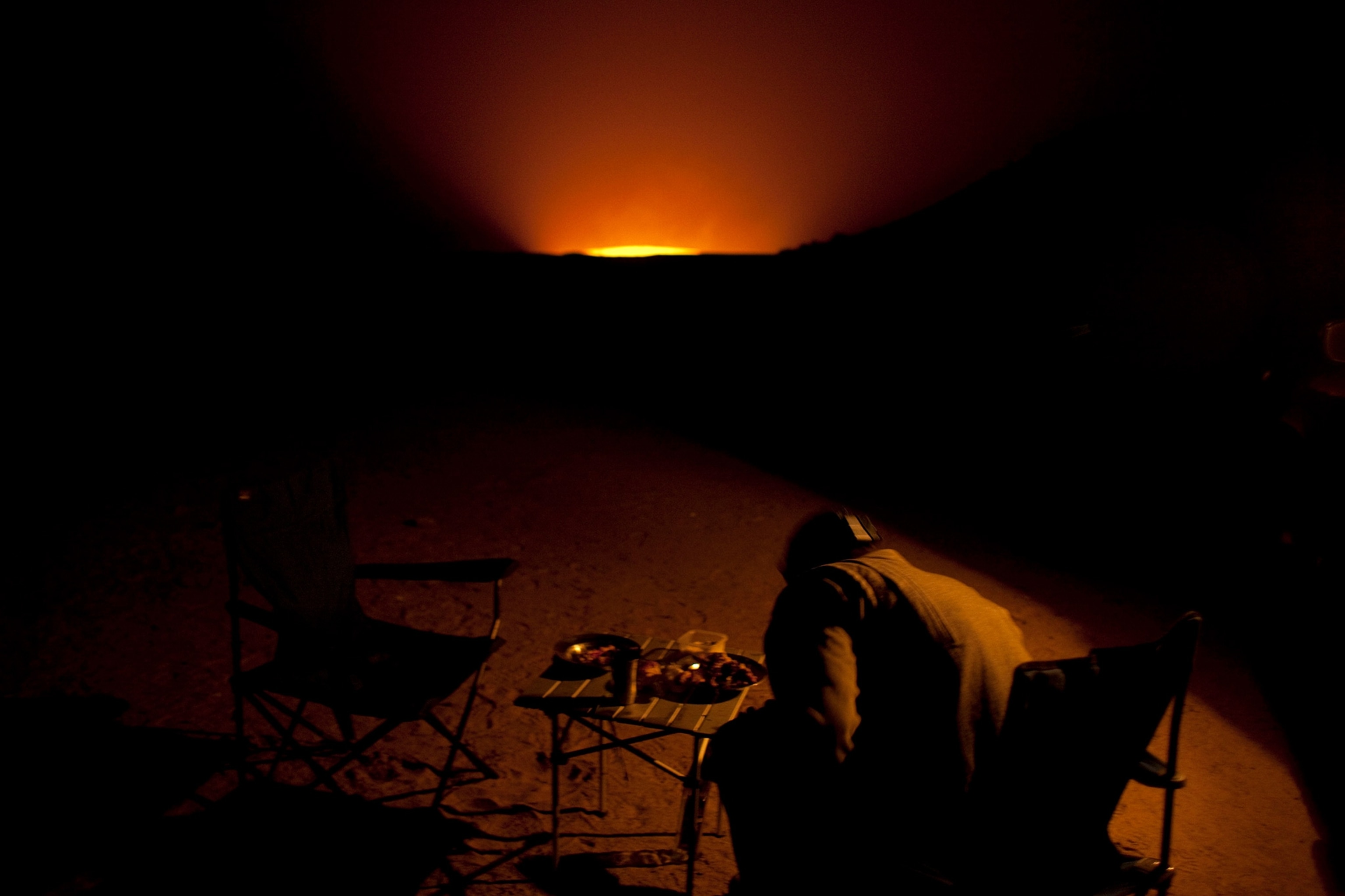 A Russian tour guide sits at a camp site near the Darvaza gas crater, glowing in the distance, at night.