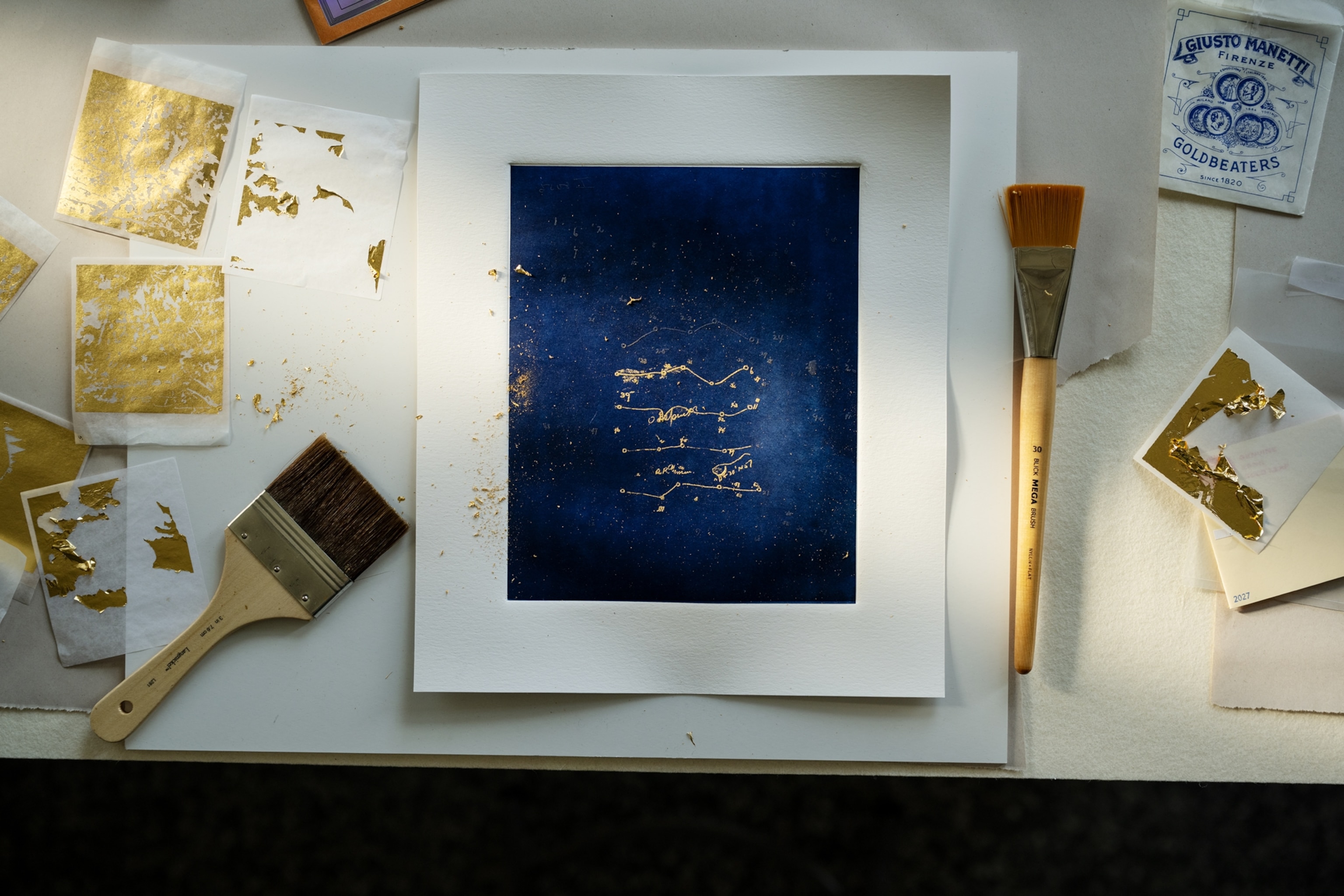 Picture of desk with dark blue print, brushes, and gold leaf for gilding.