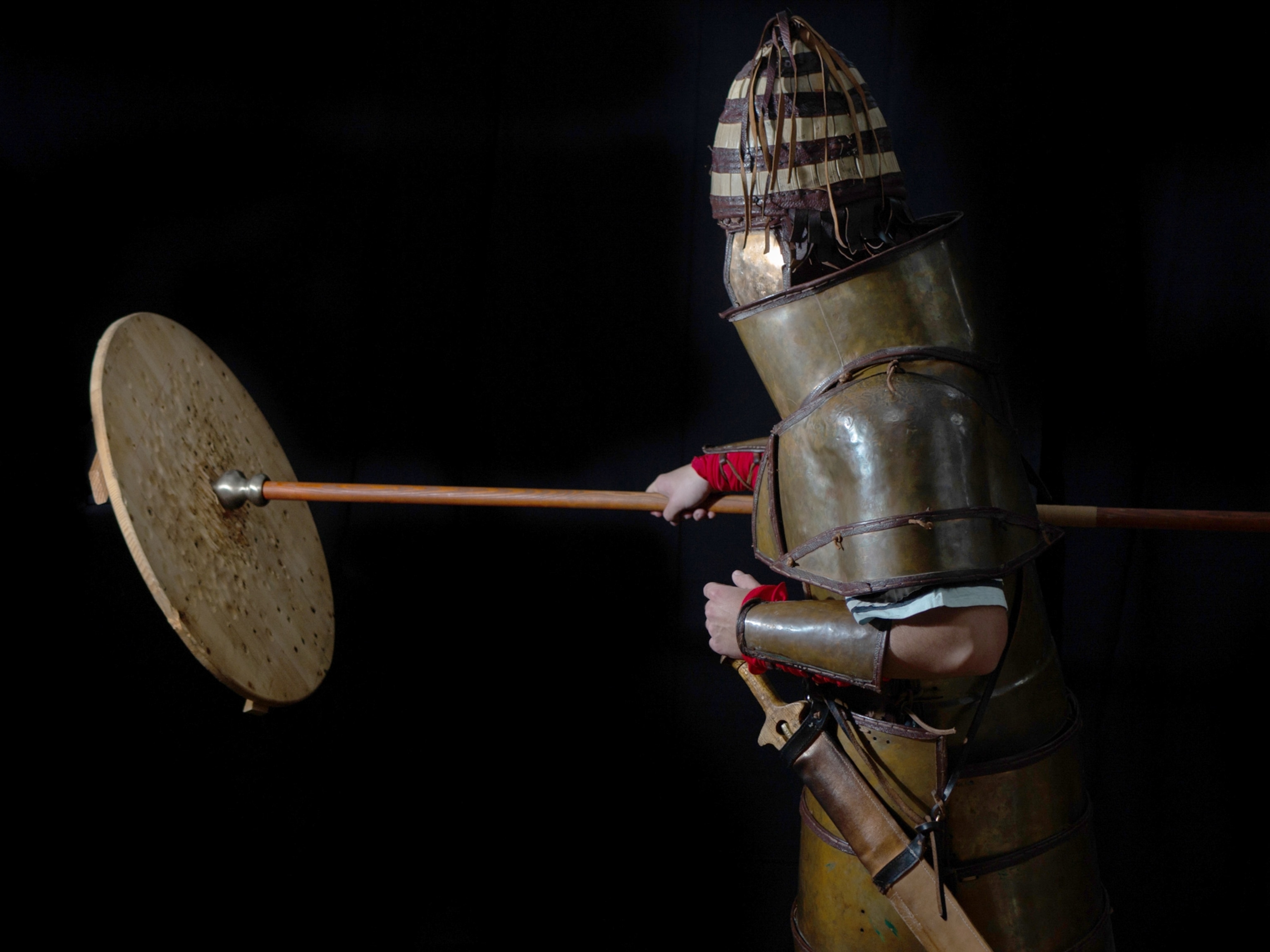 A persone simulates hitting a shield with a spear while wearing armor.
