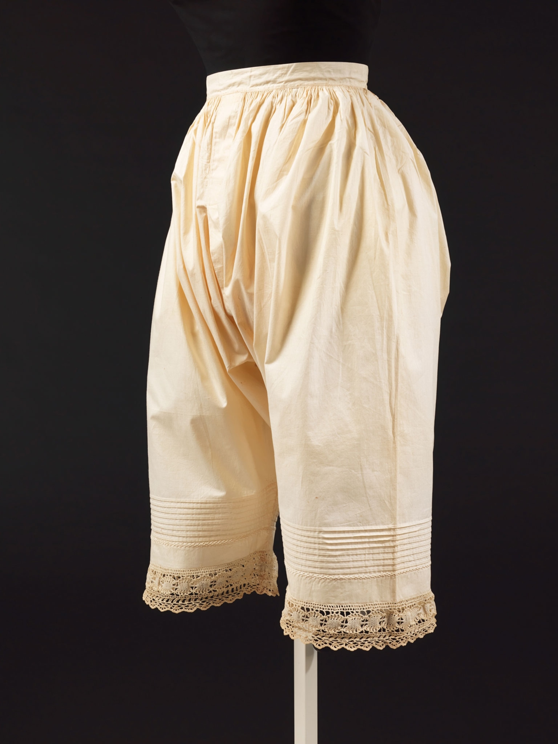 A pair of long, cream pants that are sinched at the waist, balloon around the bottom that stop just below the knee against a black background.
