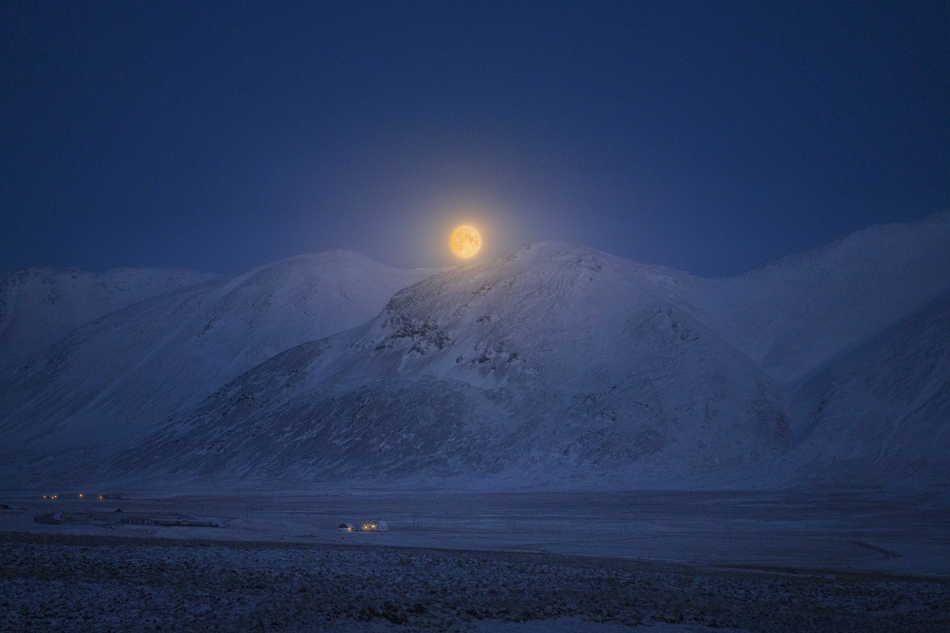 A full moon rises over Iceland's winter-clad mountains, illuminating the stark beauty of the snow-draped landscape.