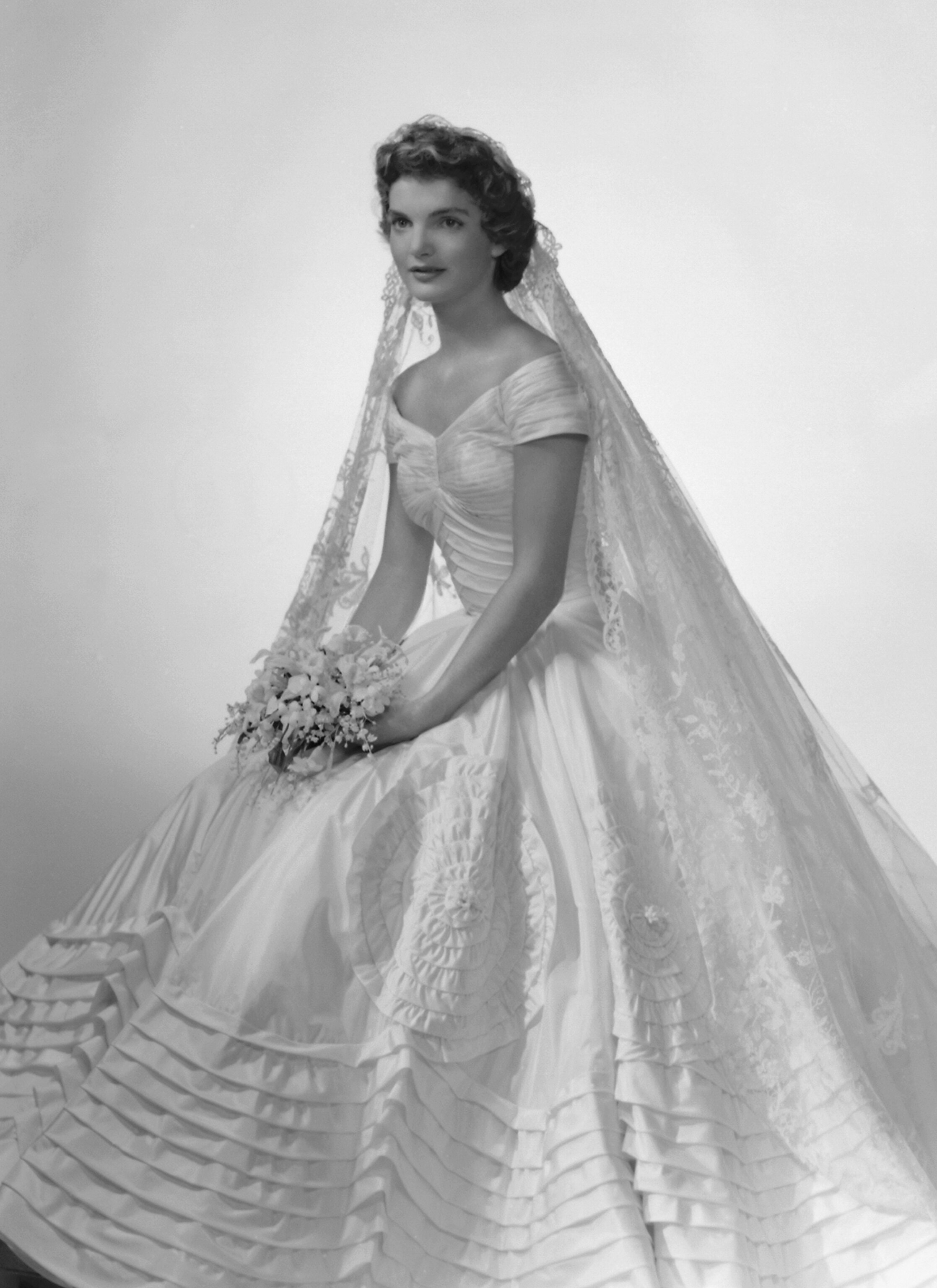 Bridal portrait of Jacqueline Lee Bouvier shows her in an Anne Lowe-designed wedding dress and a bouquet of flowers in her hands.
