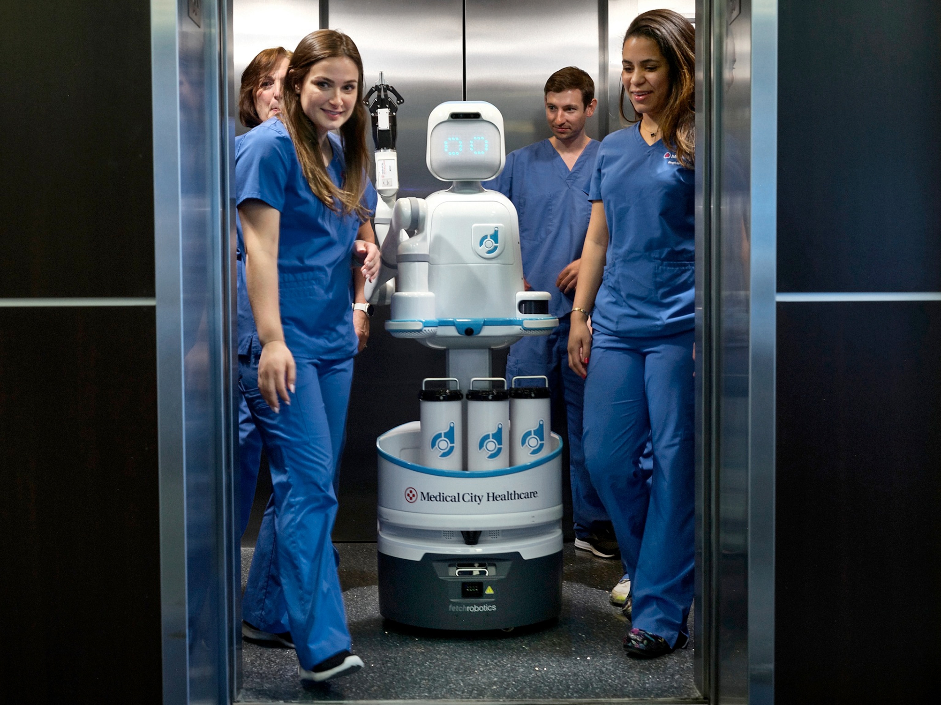 nurses in blue garbs exiting an elevator with white robot on whiles.