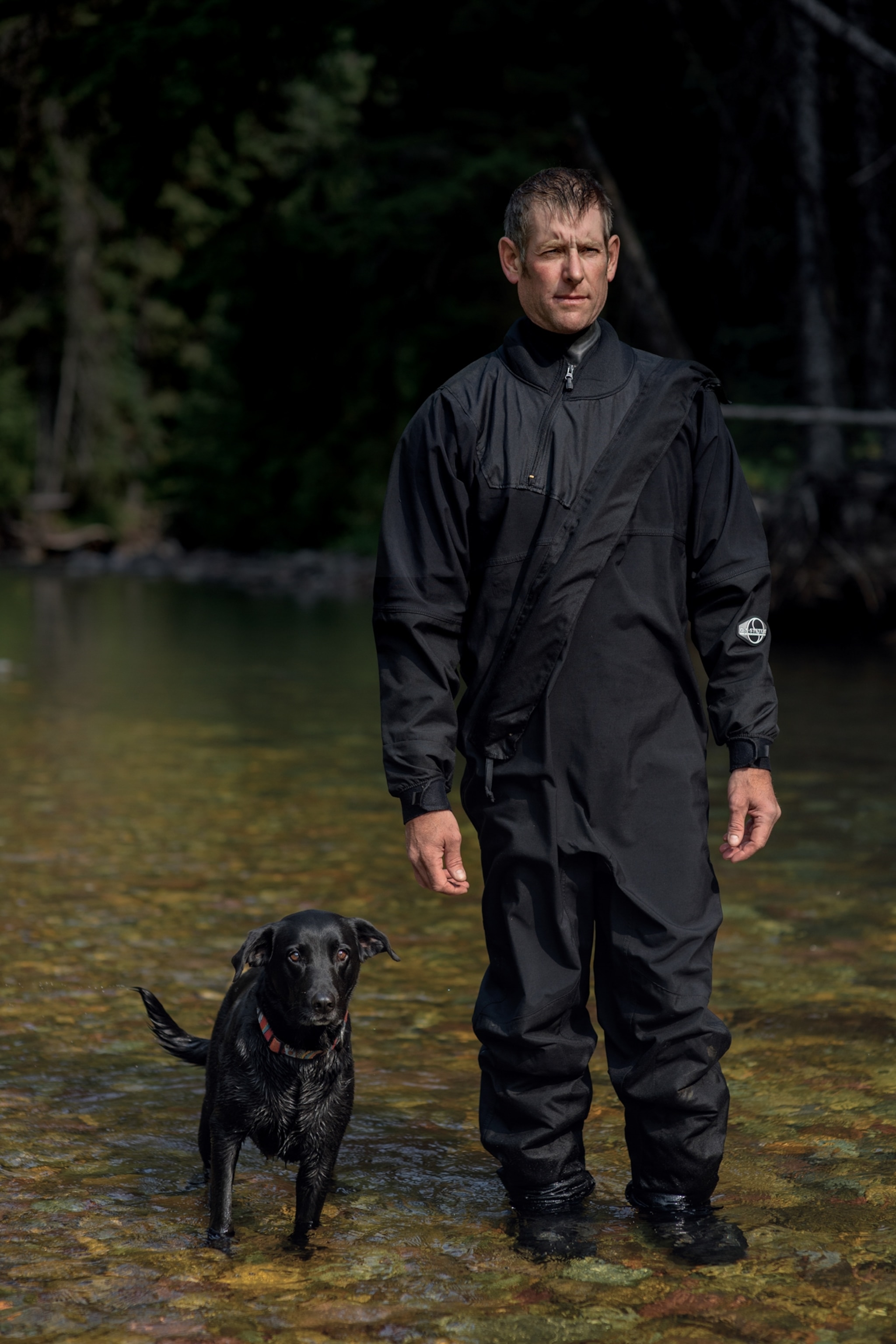 Picture of Clint Muhlfeld, dressed all in black, standing for a portrait beside his black dog.