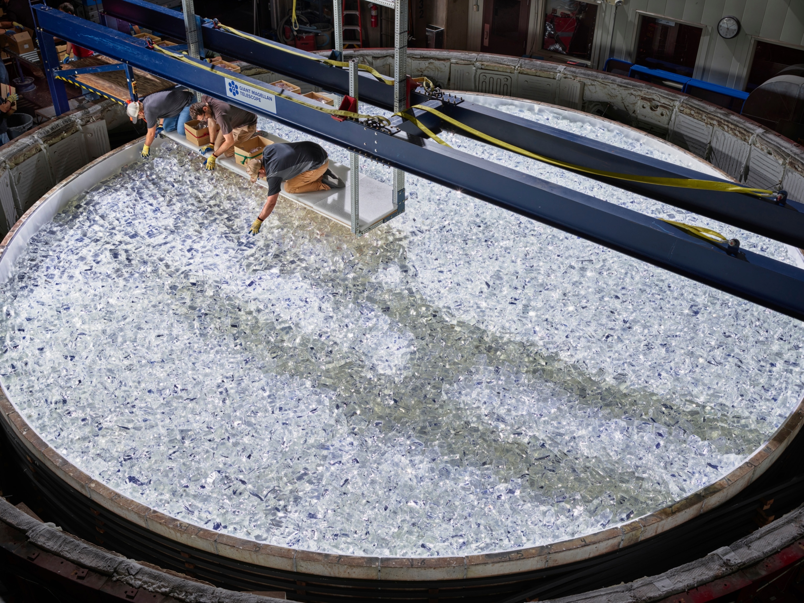 three individuals are seen crouching on a raised platform over a massive vat of crushed glass.