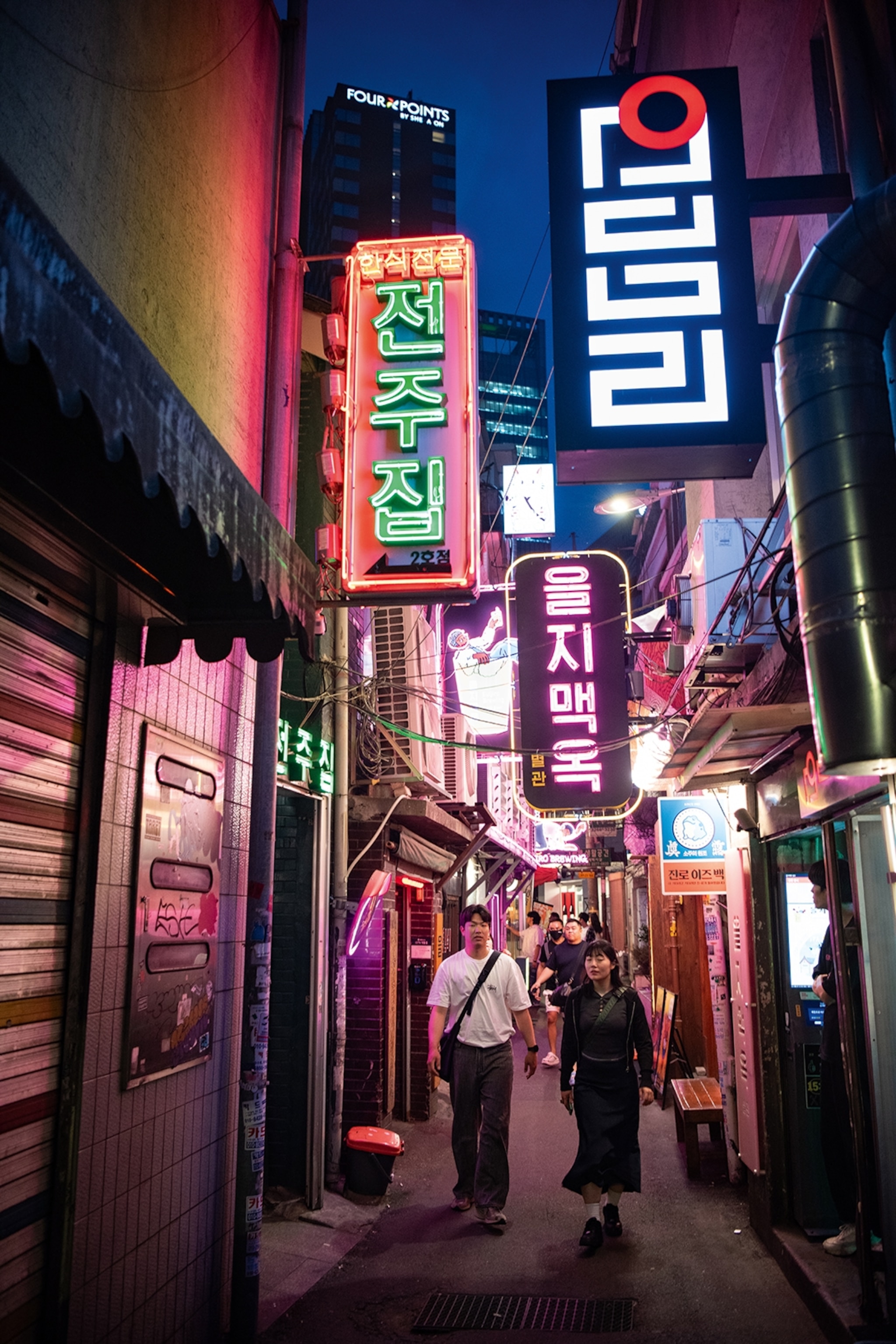 A man and woman walk down a narrow inner-city streetwith bright neon signs hanging above them