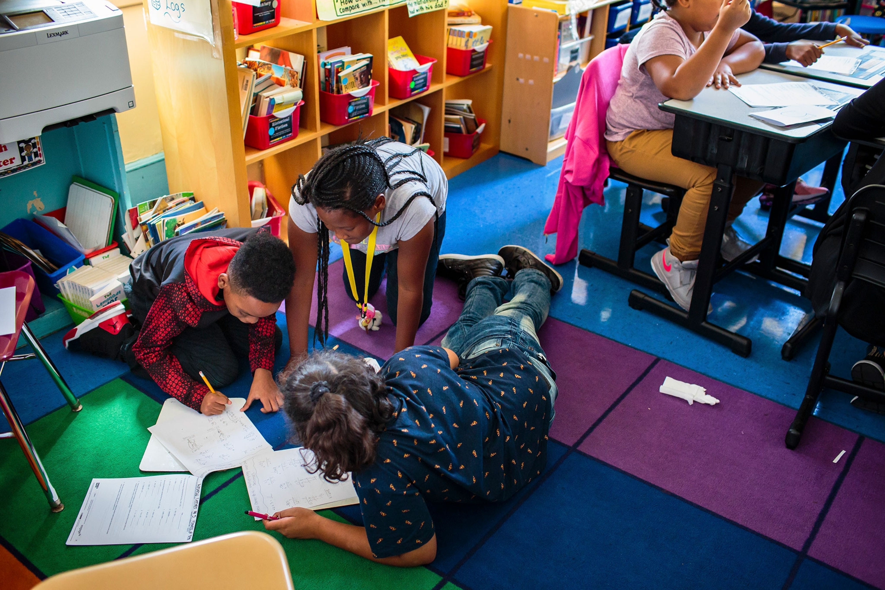 Students work on schoolwork on while sitting on the floor covered in colorful carpet and surrounded by school supplies and equipment.