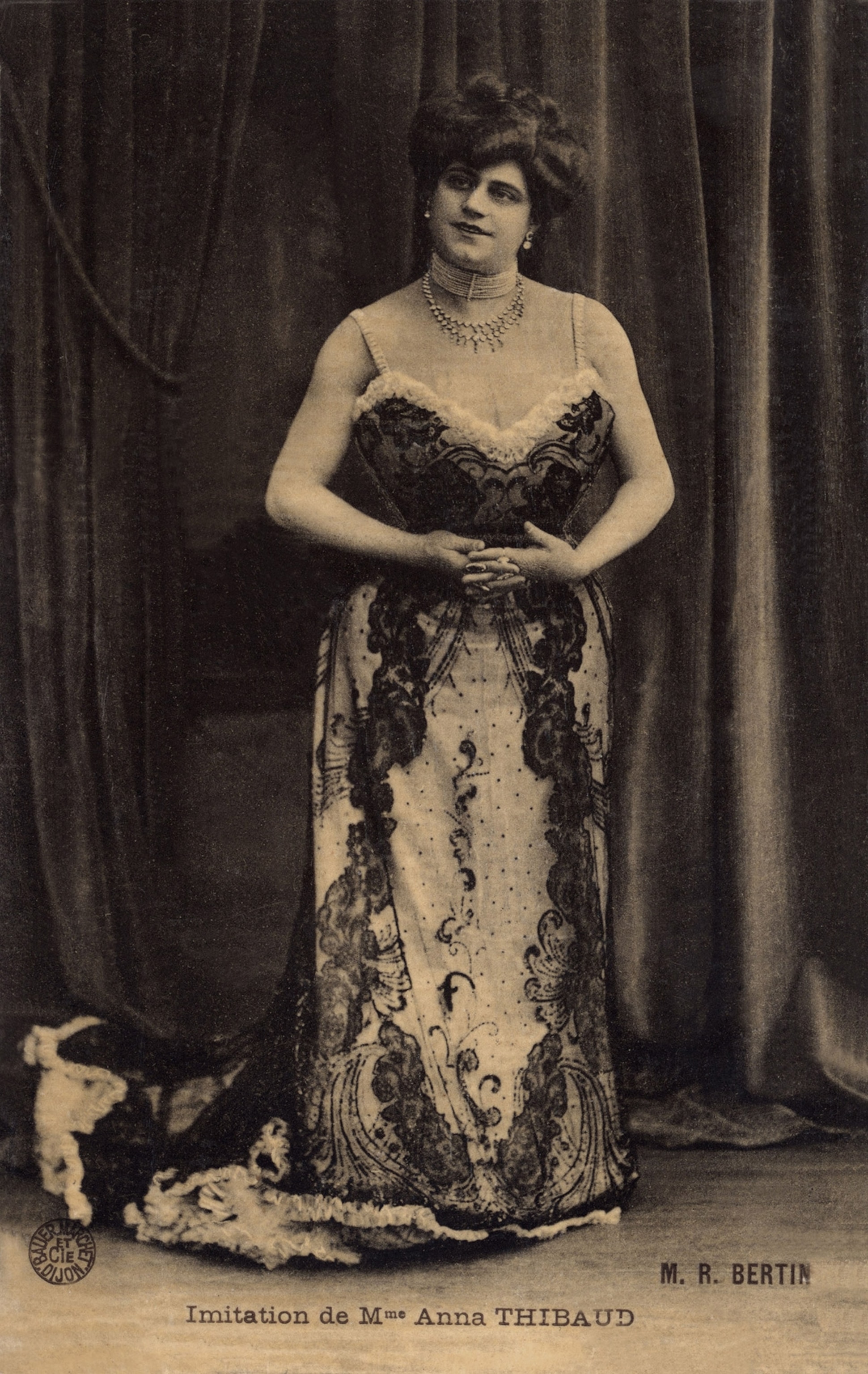 A speia toned image a woman stands hands clasped in front of a heavy curtain dressed in an ornate gown.