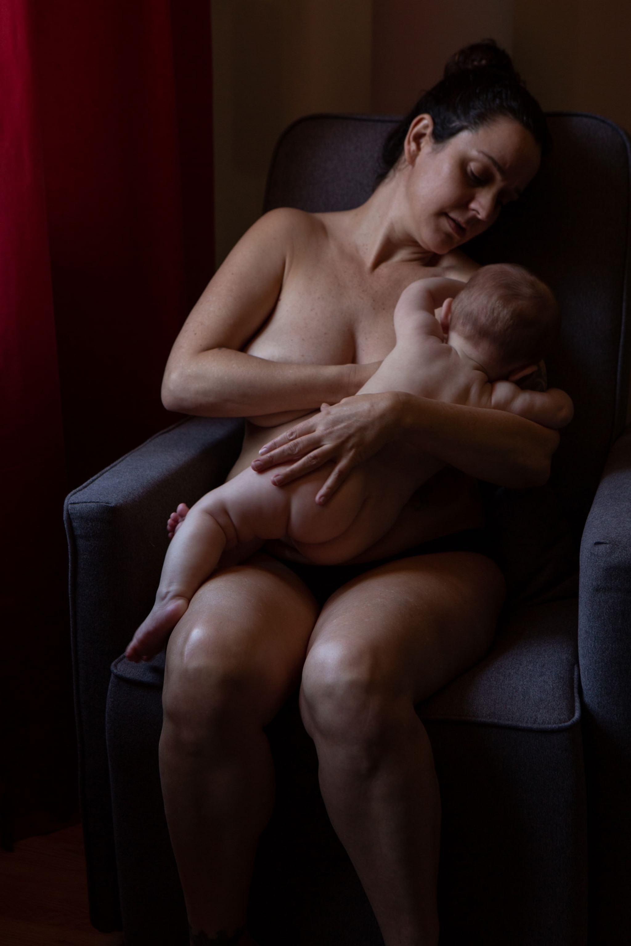 The mother holds the baby at her breast in a chair.