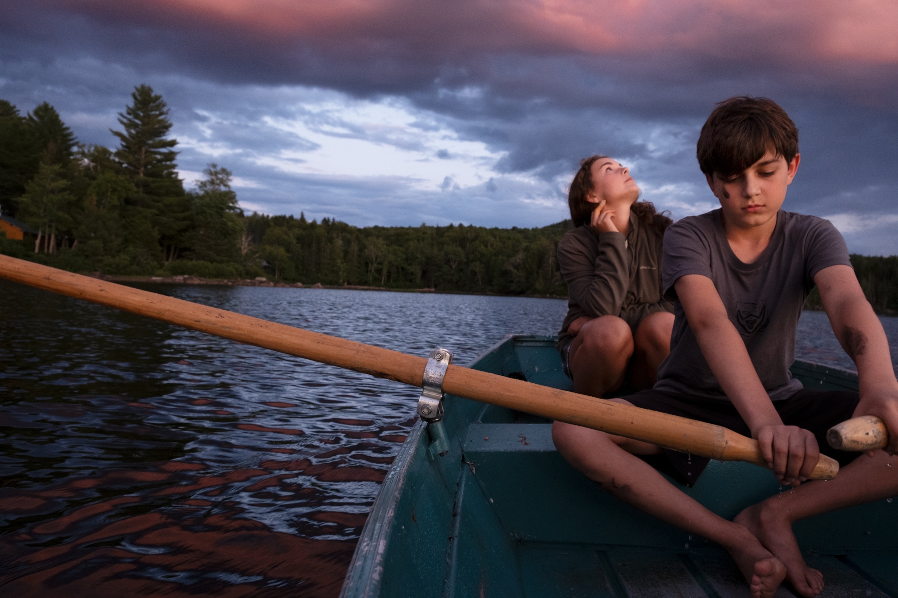 Two kids pictured sitting in a canoe on a lake at sunset. The one in front is paddling, while the one behind looks to the sky.