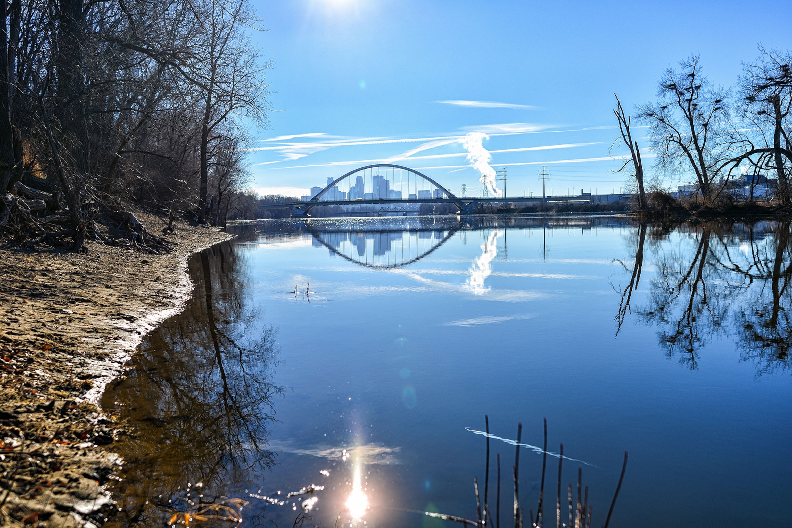 A view along the river bank of the Mississippi River. Instead of snow and ice, the bank is muddy and the water open. The skyline of Minneapolis can be seen in the background.