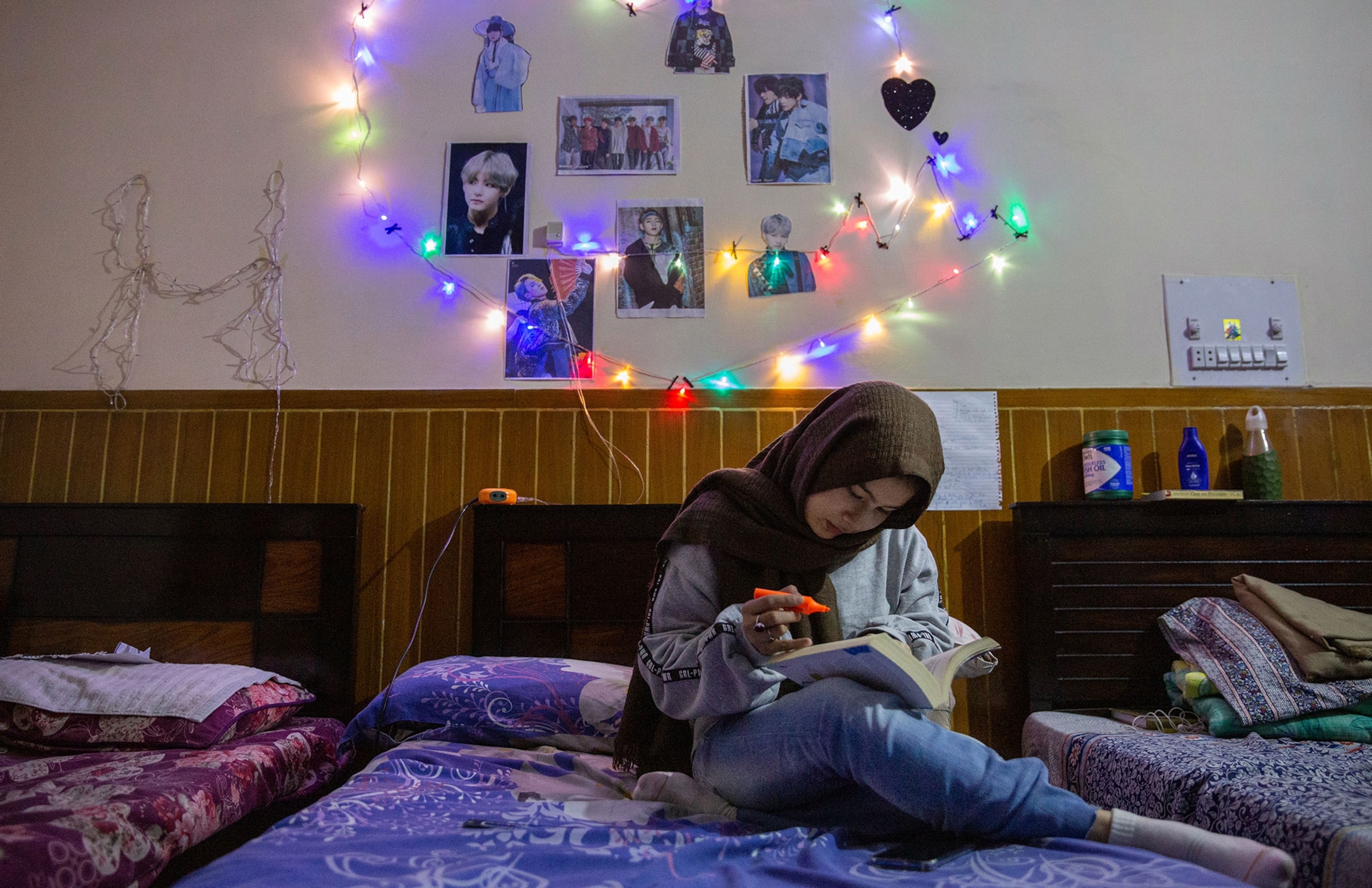a woman sits on her bed looking at a textbook, multicolored lights hang above the headboard
