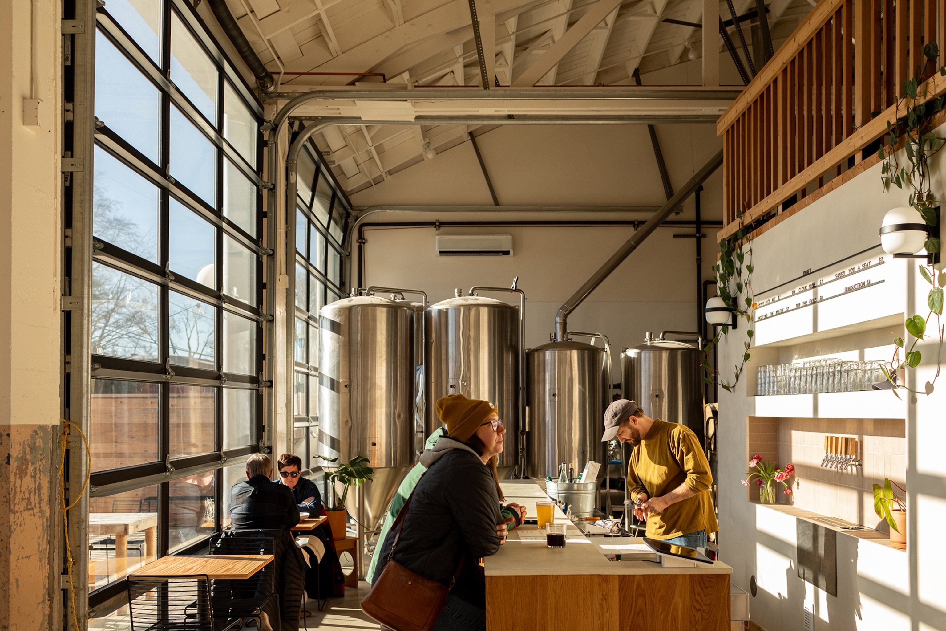 Patrons sitting in a brewery taproom. Metal tanks line the back wall, whilst the sun is streaming in through the window on the left.