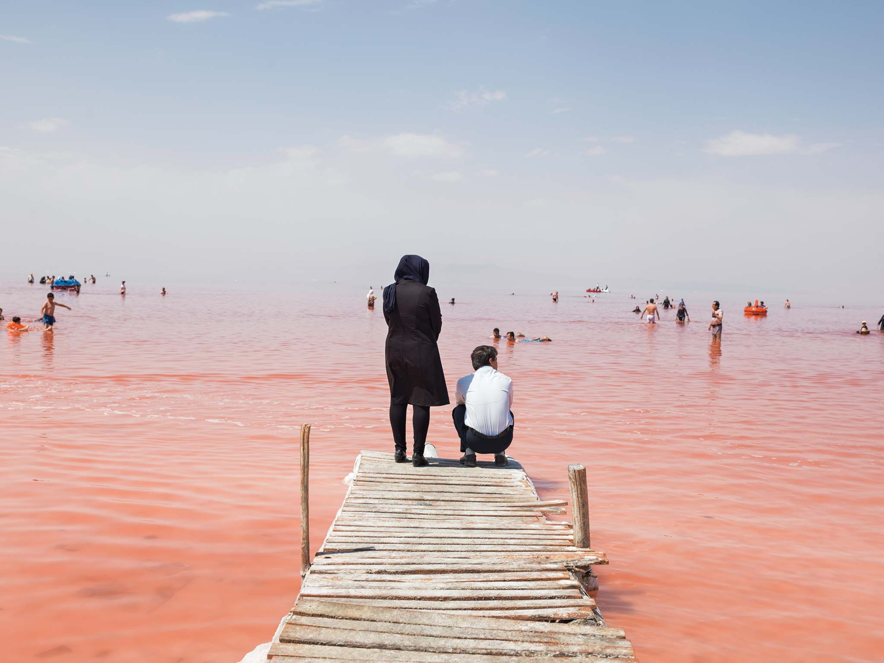 A woman stands and a man squats on a dock over a reddish pink lake