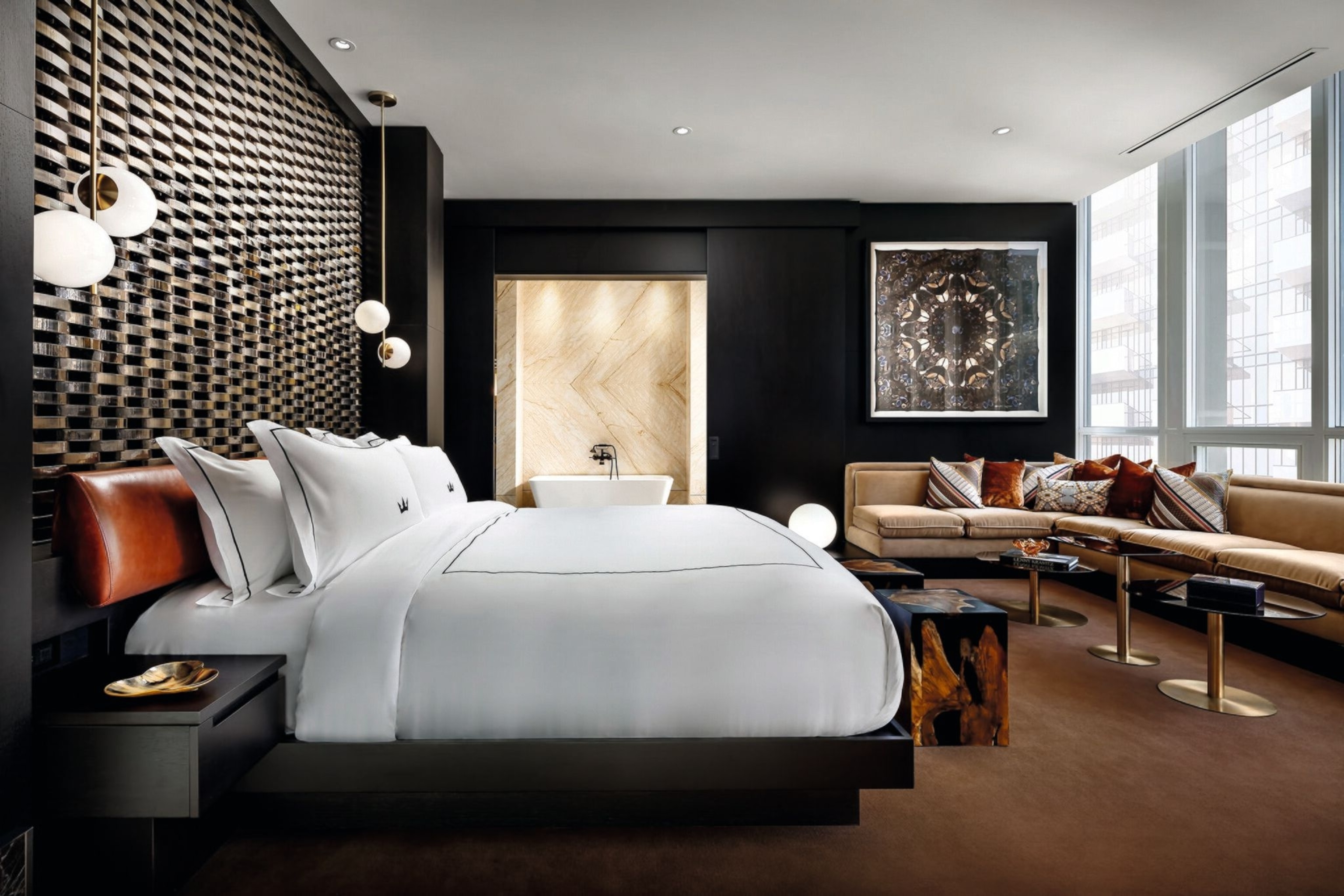 A large bed with white sheets. Opposite, a room-long sofa sits below a wall of windows.