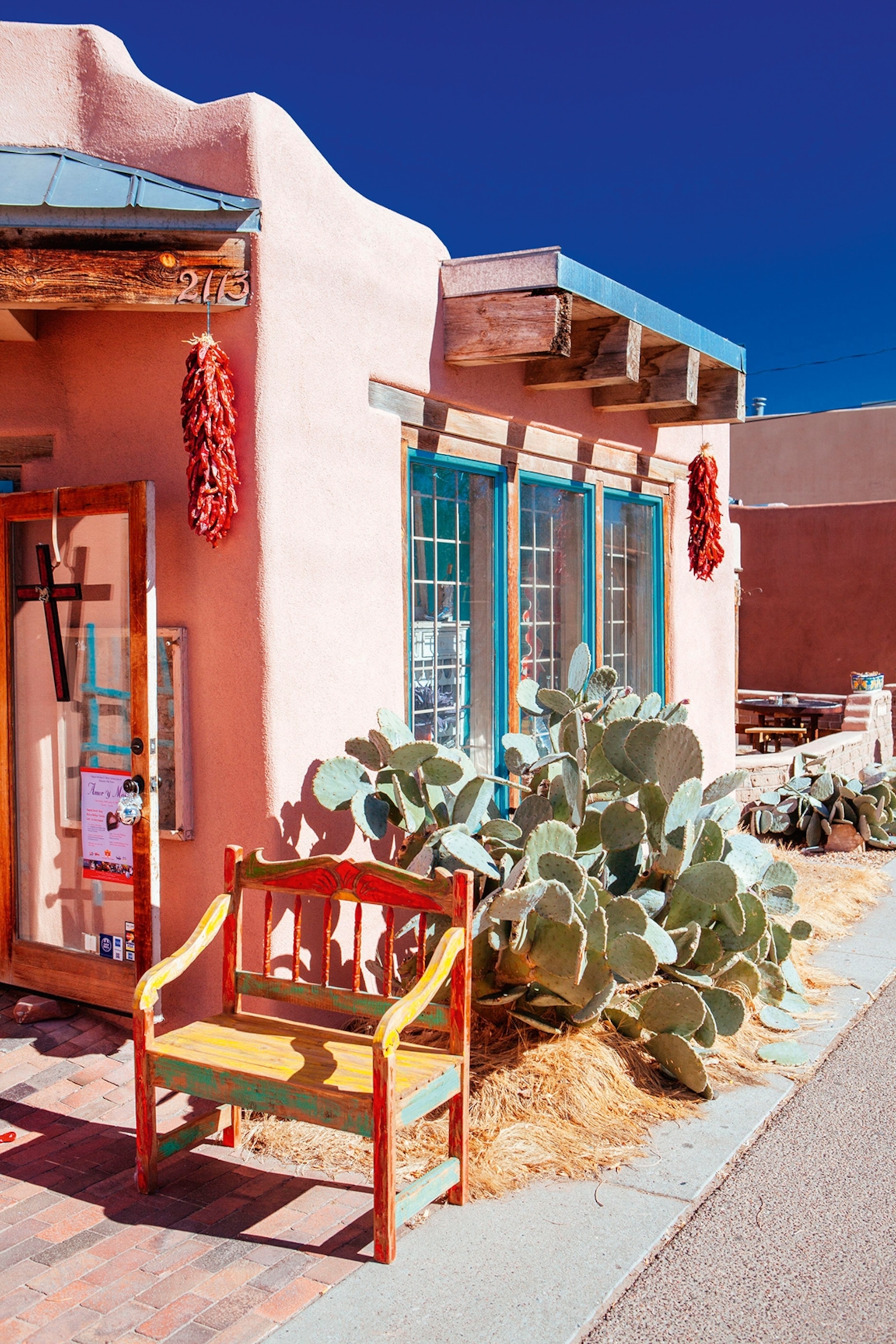A colourful, round-edged sandstone house with bunches of dried chilis hanging off wooden beams and a bench and cacti out the front.