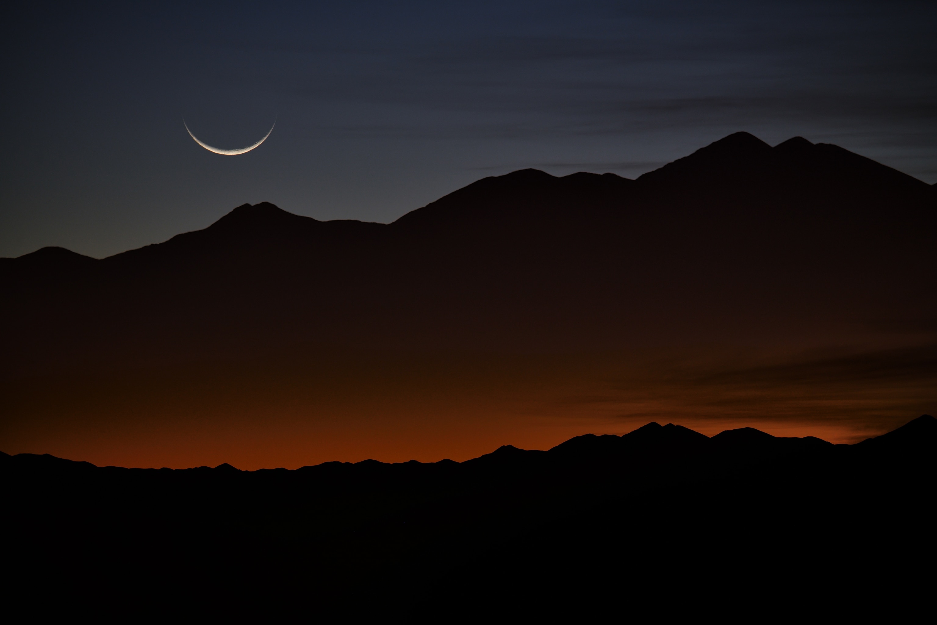 A crescent moon setting at sunrise over the mountains