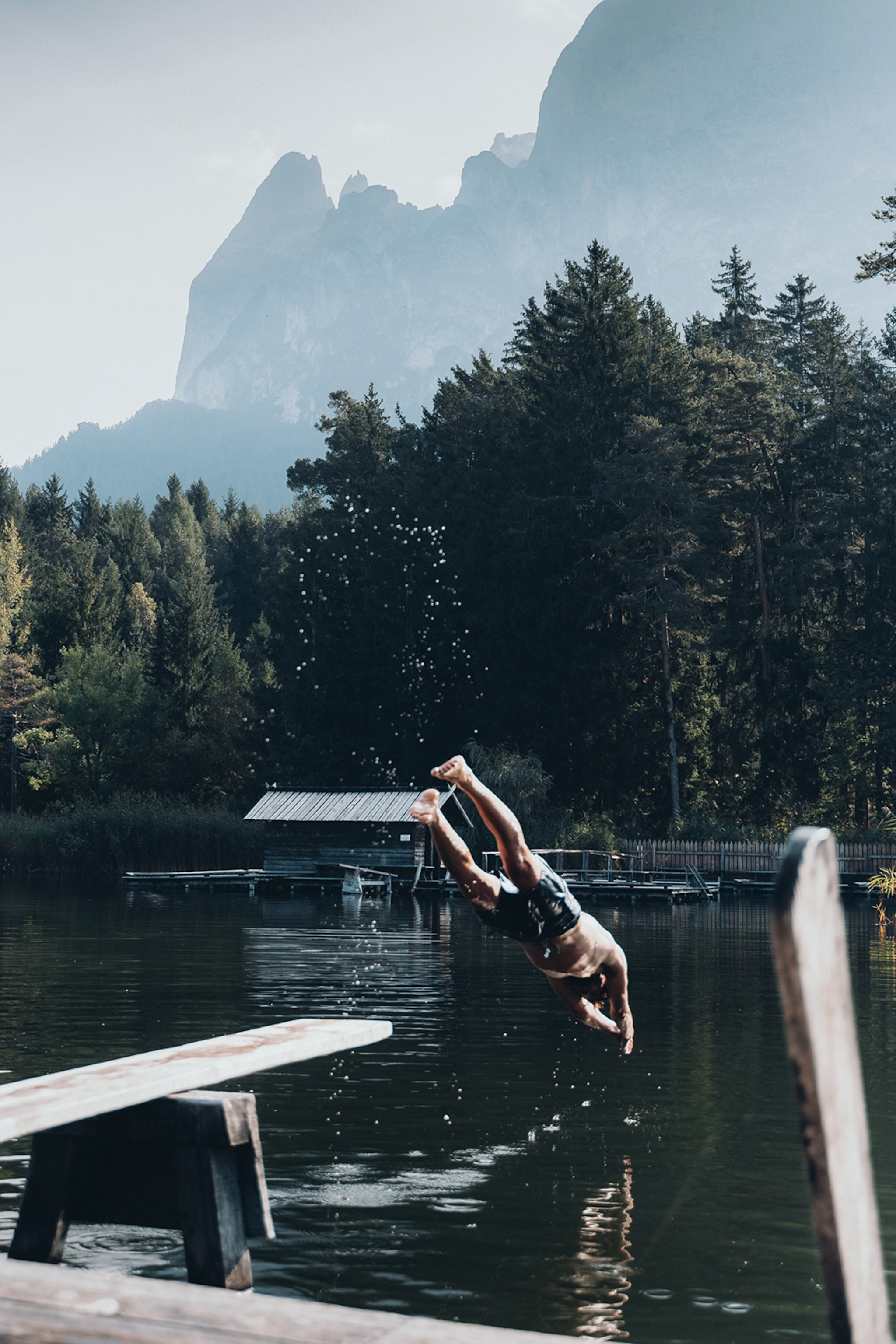 A person diving from the bank to a calm lake in the dolomites, tall birch trees line the edge of the lake whilst mountains tower over in the background. .