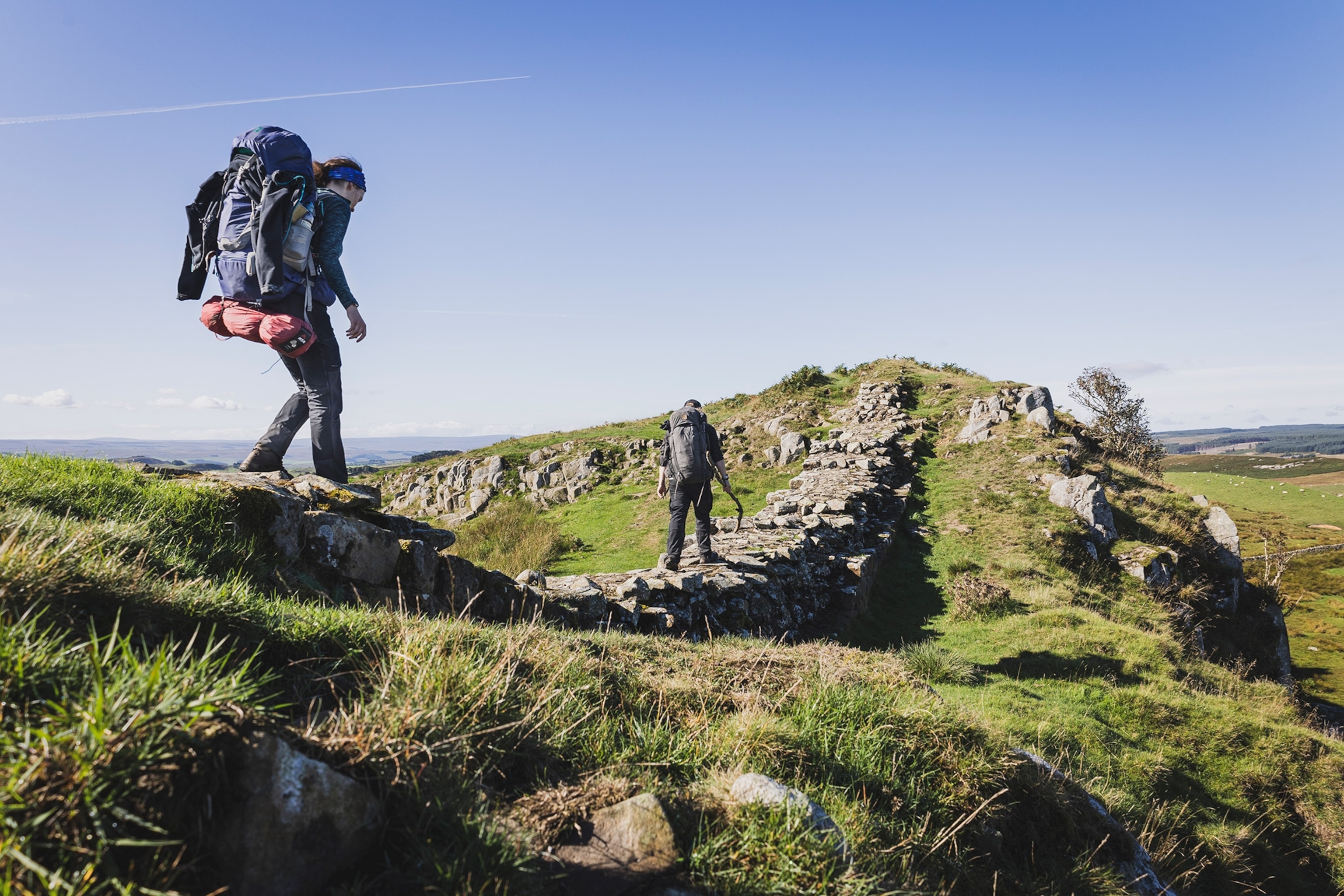 Two people with backpacks walk along the stones of Hadrian's Wall at Windsill Peaks