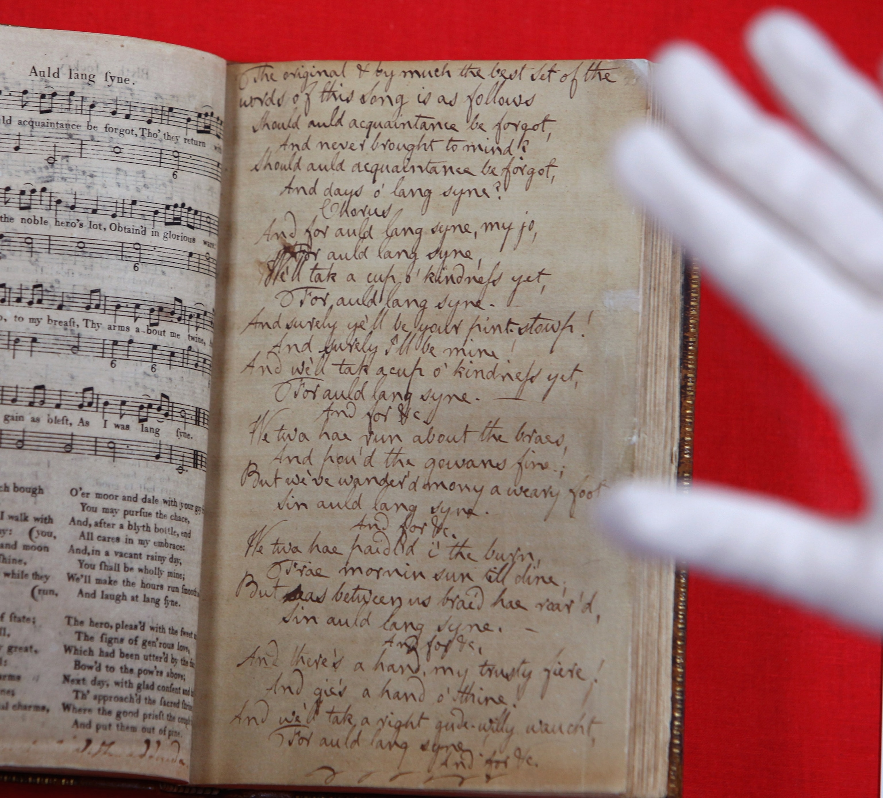 Photograph of Robert Burns manuscript and lyrics to Auld Lang Syne against a red background. A hand hovers over the right side of the image.