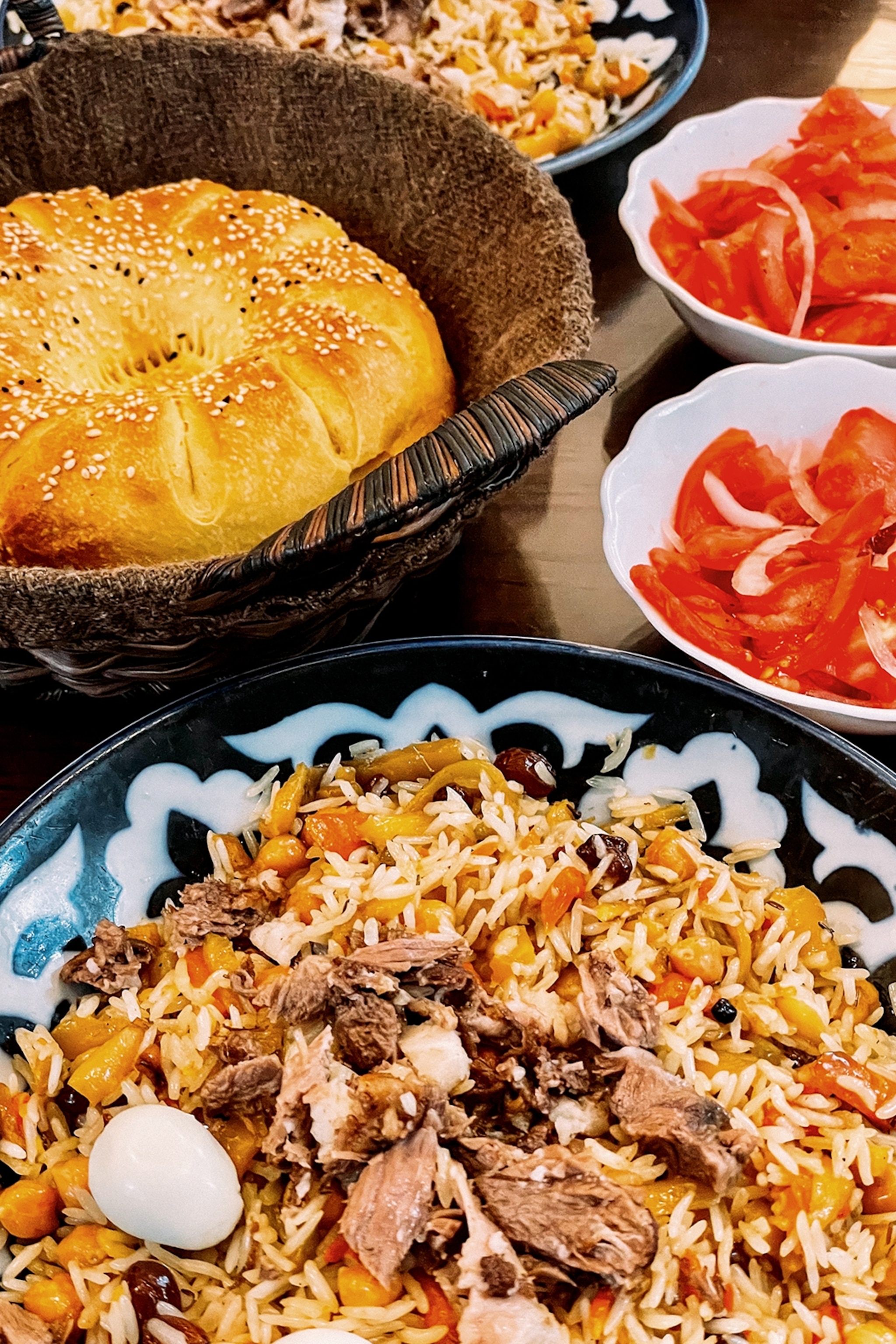 A set table with bowls of tomato salad, sesame-strewn bread and a rice dish with pulled meat, raisins and vegetables.