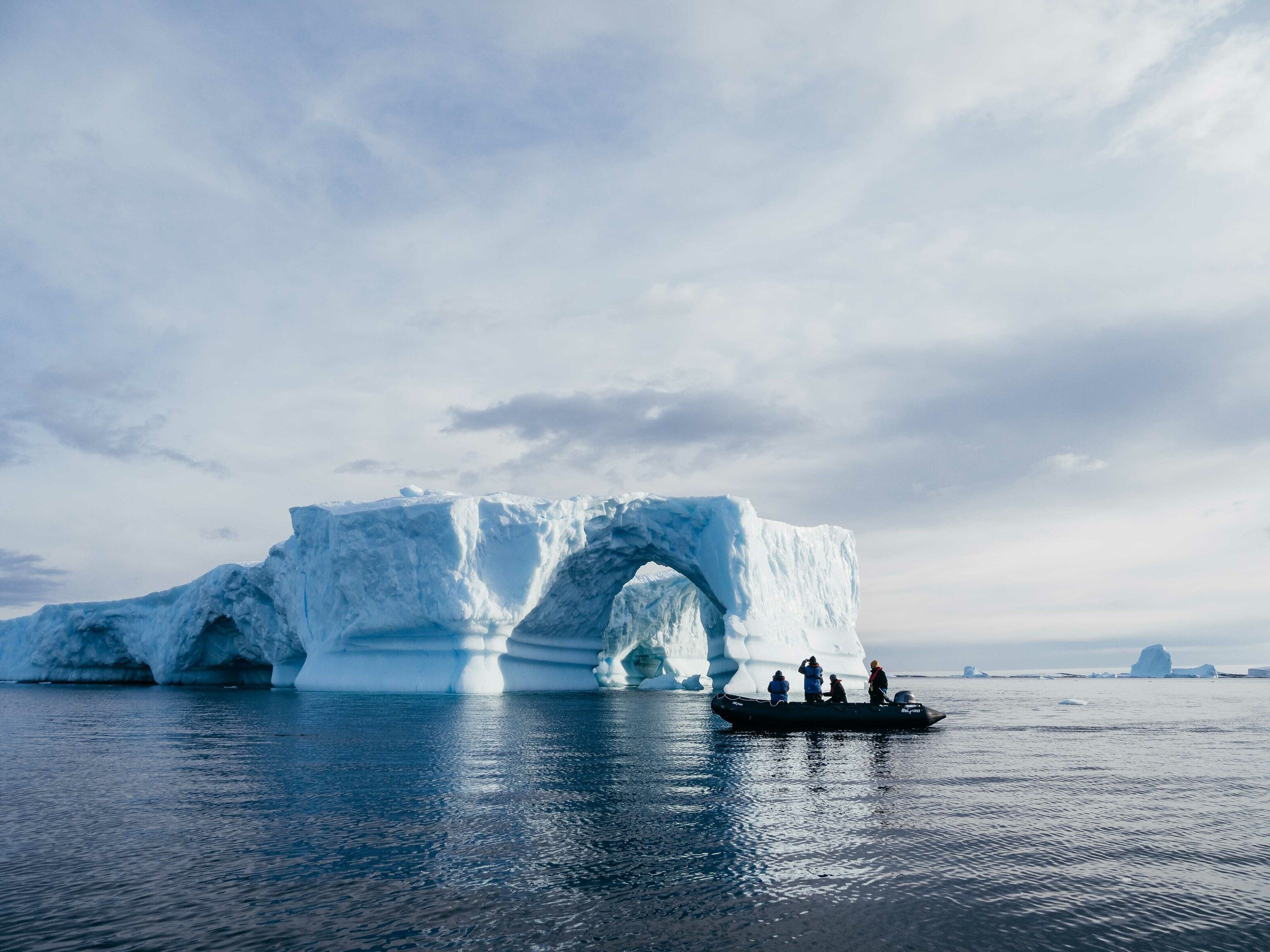 Four people in a small boat looking at an iceberg, which has formed a natural structure that looks more like an ice bridge between two large white rocks.