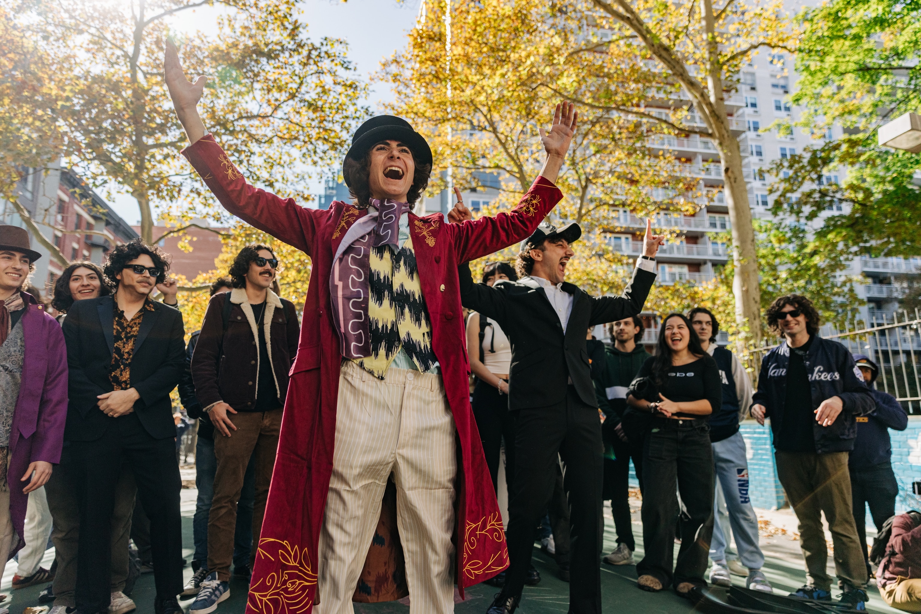 A young white man dressed in a top hat and coattails gestures and grins outdoors in a park in front of a crowd of onlookers.