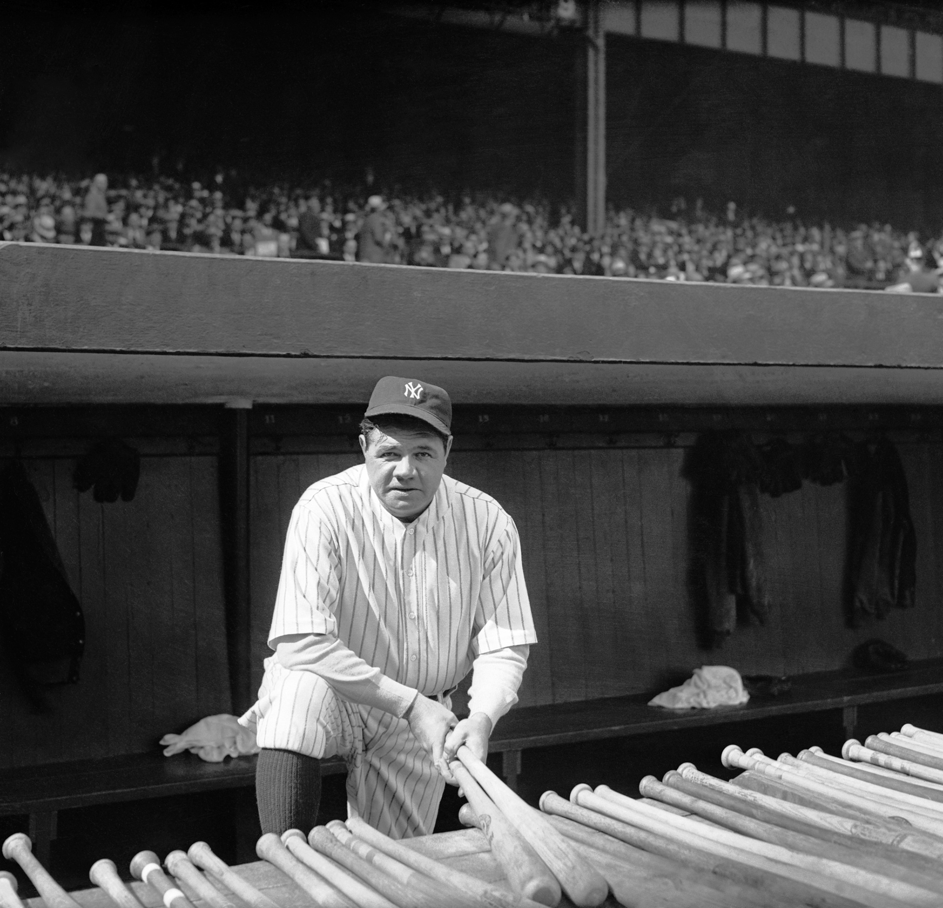 A black and white photo of Babe Ruth looking at the camera. He is kneeling forward with his foot propped up. He is in his striped uniform and there are two extra bats in front of him. There is a crowd behind him, but they are blurry.