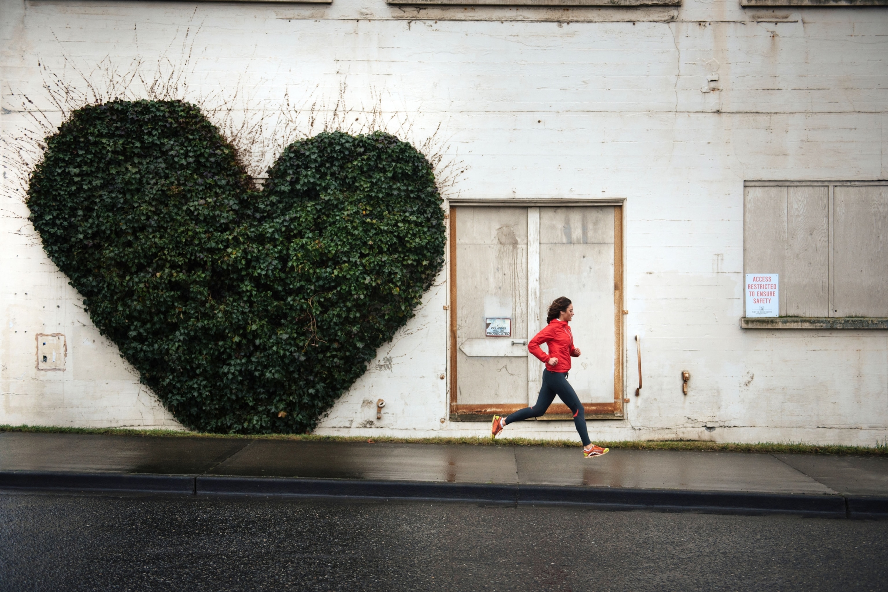 Side view of a woman in running clothes, jogging on a city sidewalk near a patch of foliage in the shape of a heart on the wall of a building.