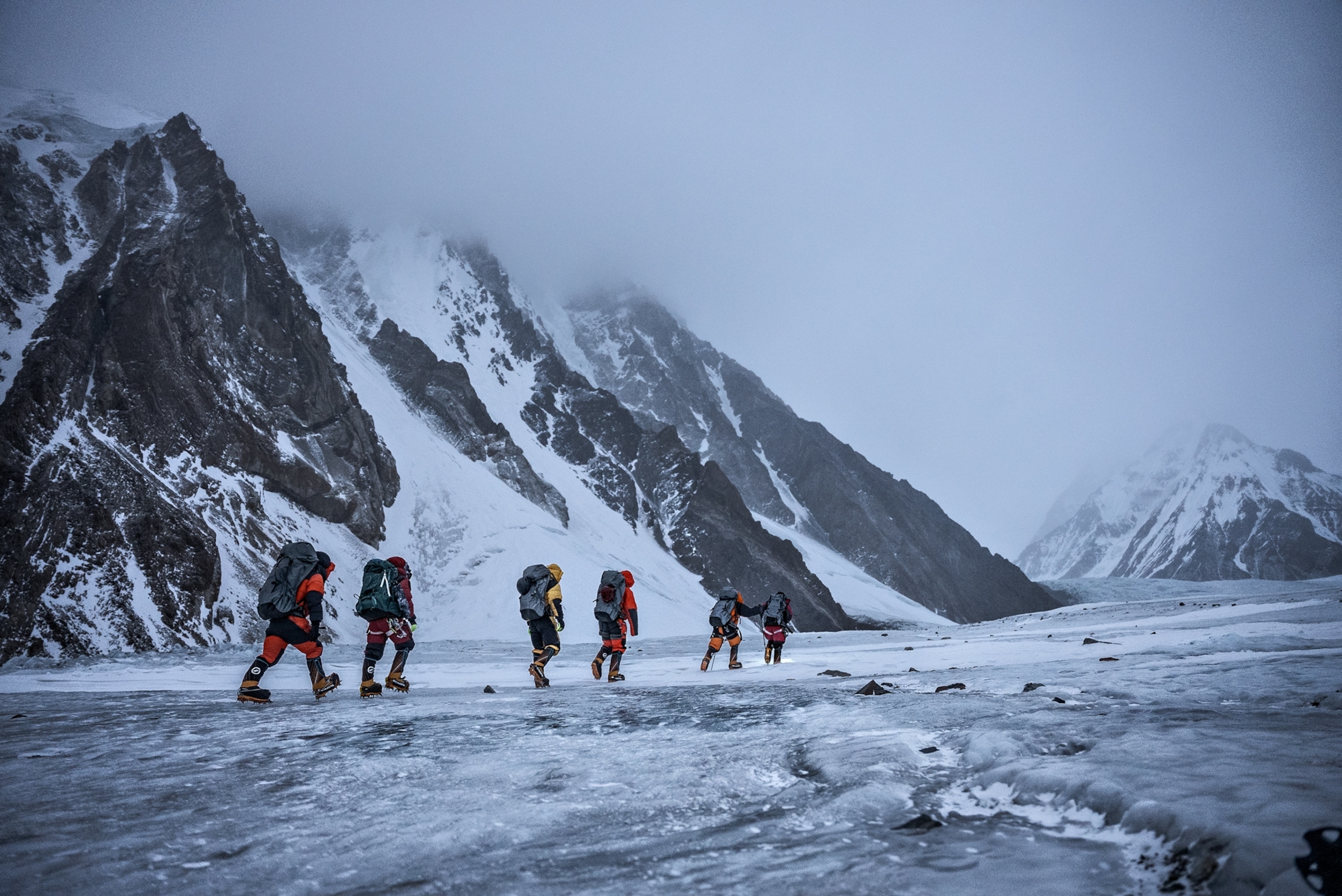 Picture of six men in crampons caring large backpacks walking on ice away from camera.