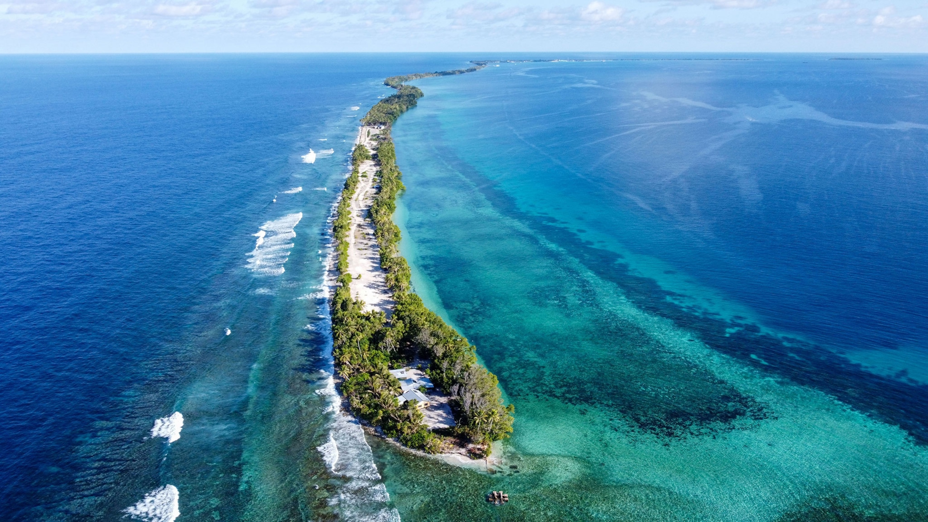 An aerial view of the southern end of Funafuti island in Tuvalu shows a small strip of land surrounded by blue water. At the tip of the island sits a house surrounded by lush green trees.
