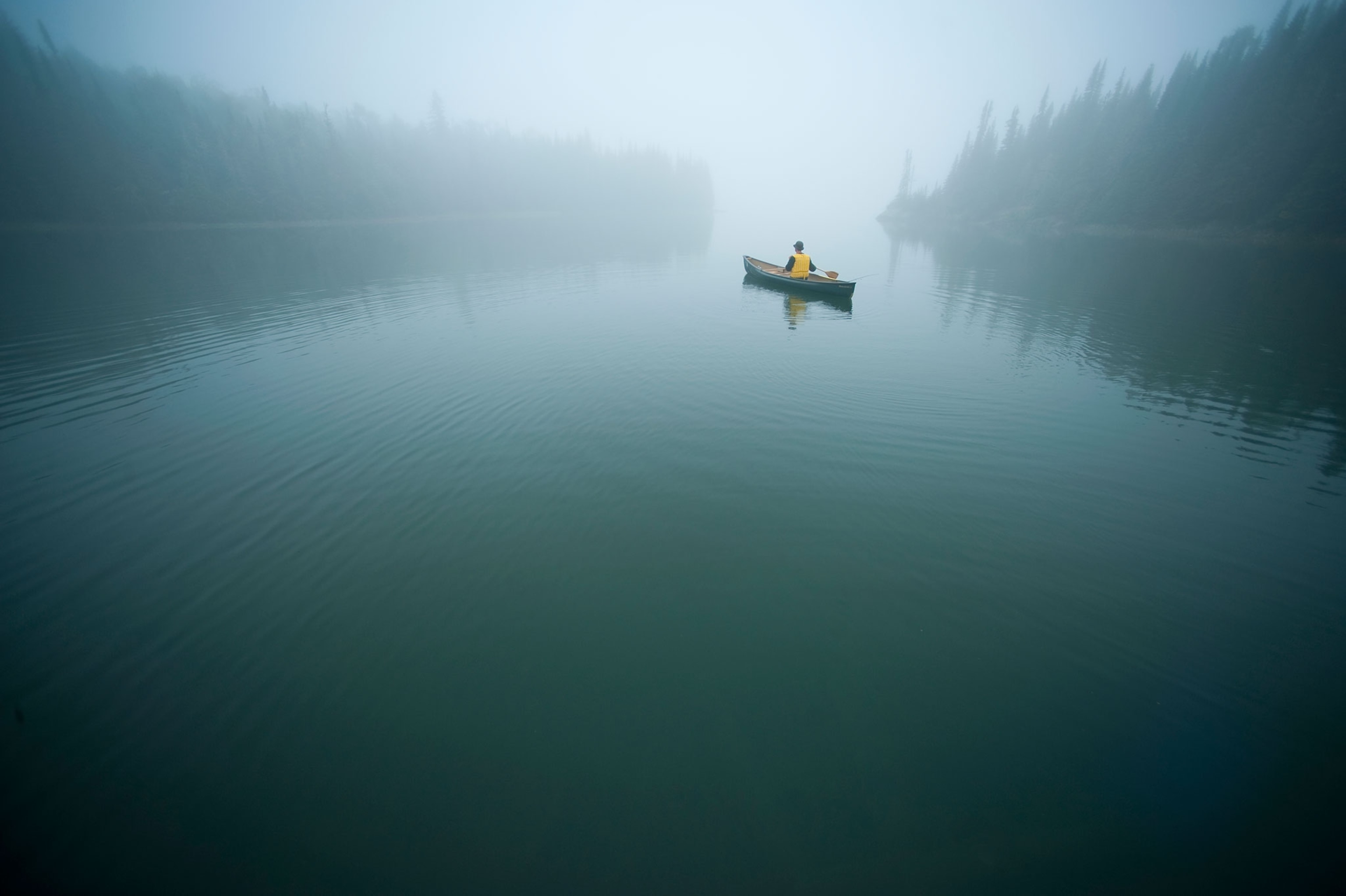 A person in a yellow life vest paddles their canoe in the middle of a foggy lake lined with trees on both sides.