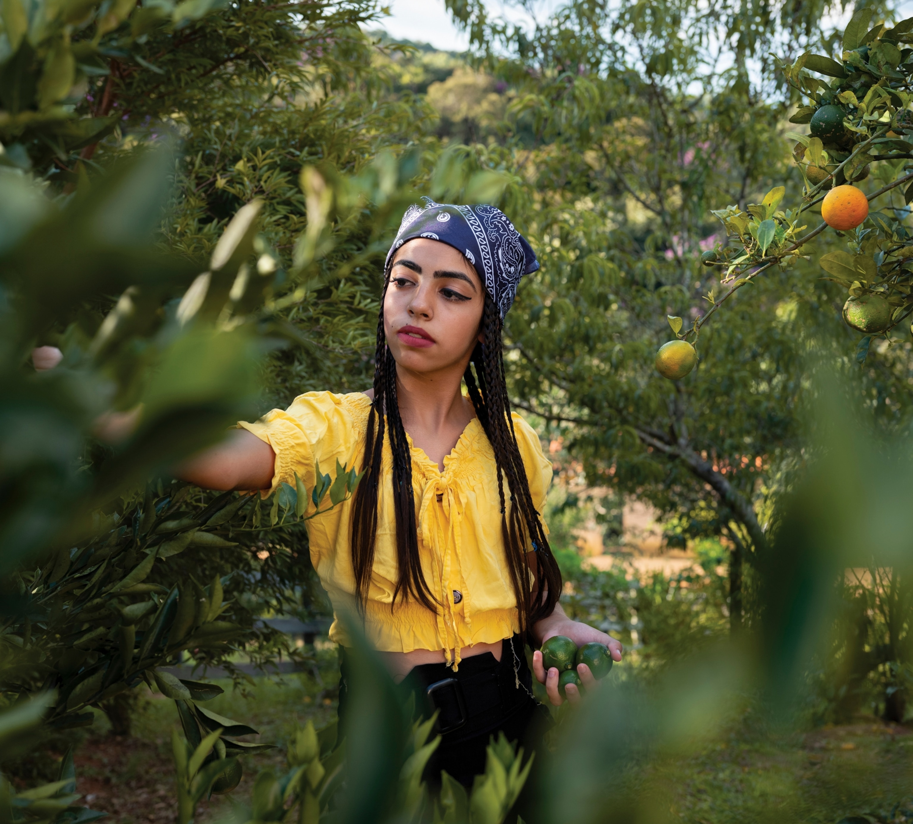A woman walks between fruit trees and reaches out to grab some off a tree. she is wearing yellow with a blue bandana on her head.