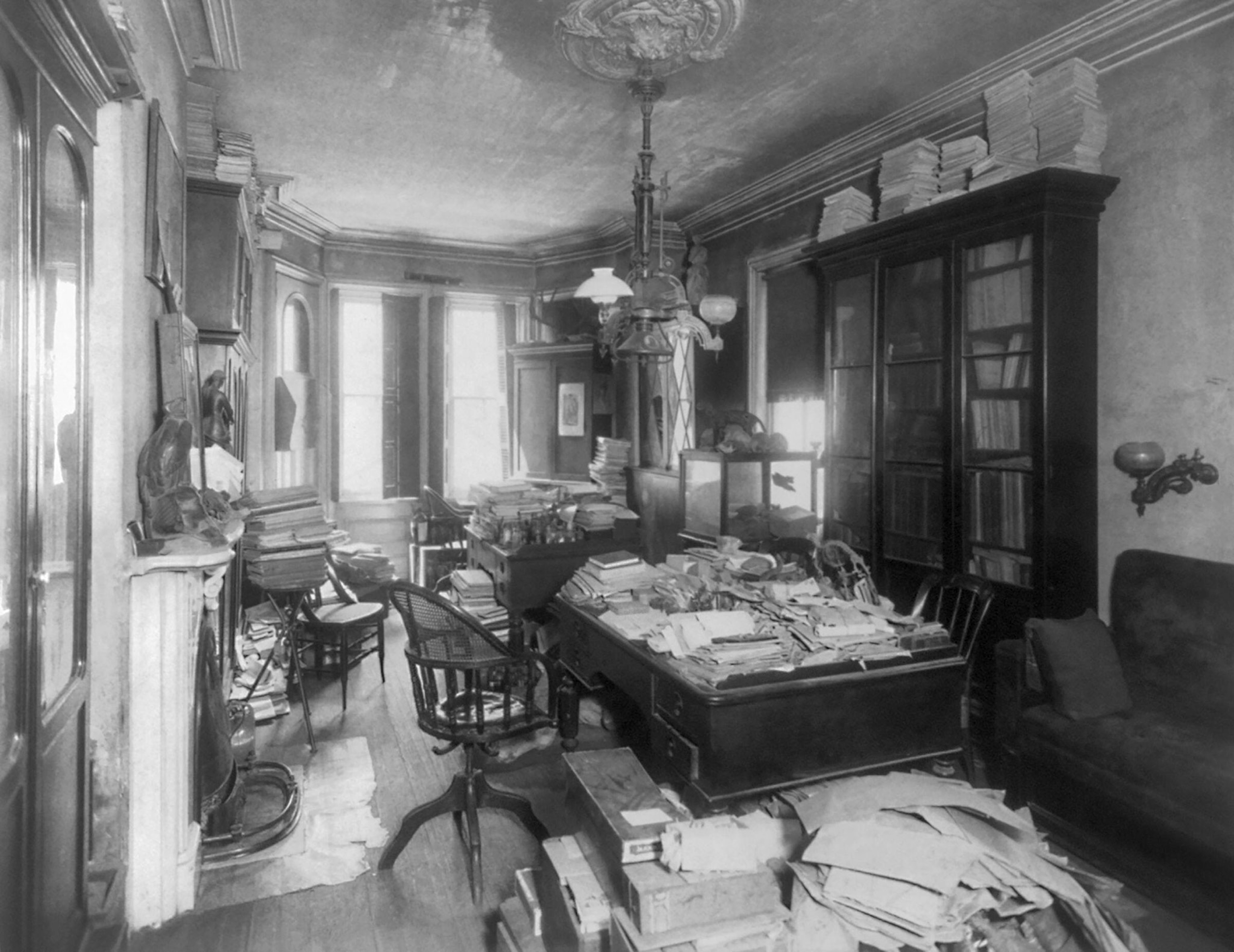 A black and white photo of an office from the late 1800s with papers and books in piles on tables, bookshelves, and the floor.