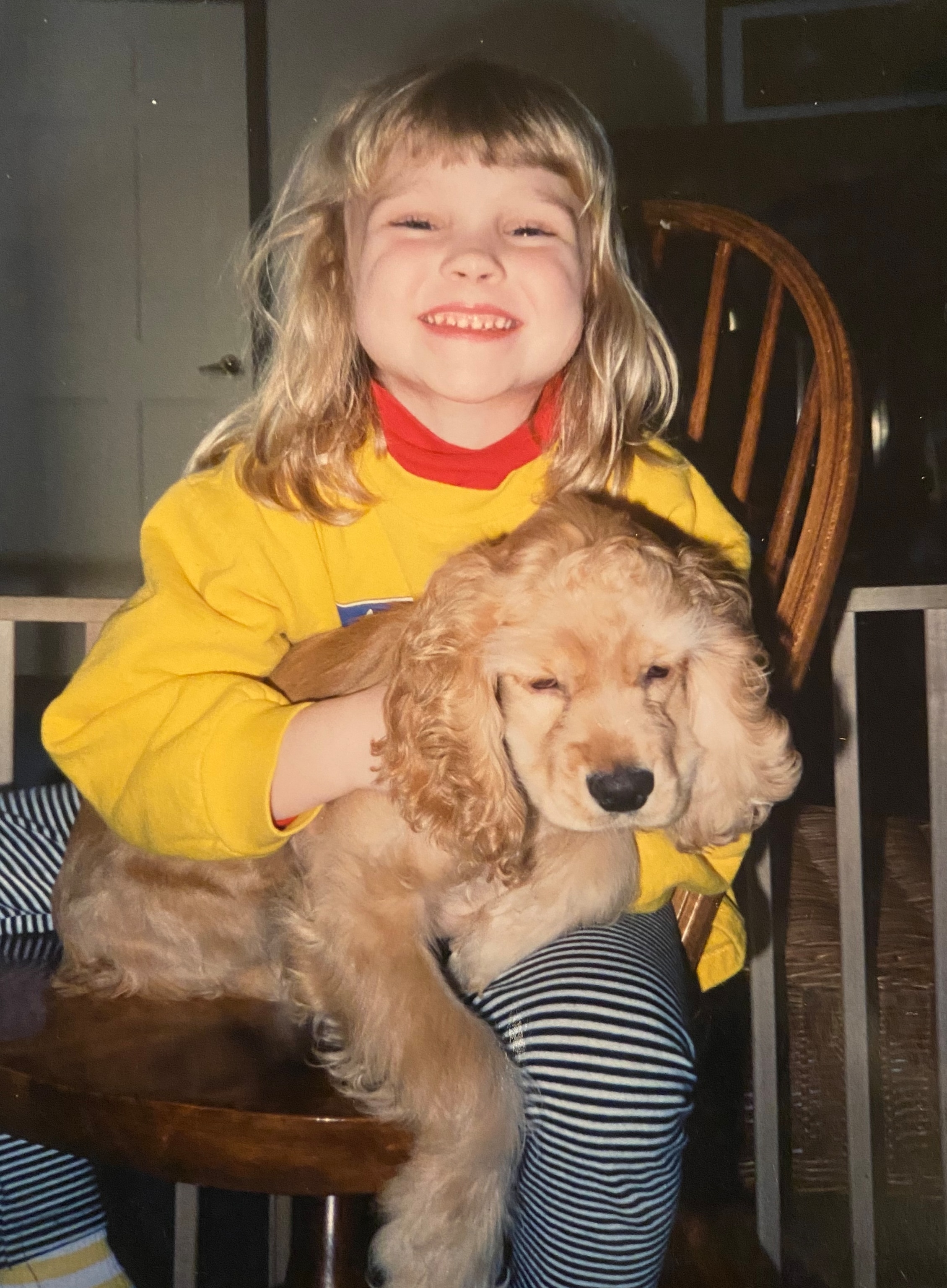 A grinning young white girl holding a dog in her lap sits in a wooden chair inside a house.