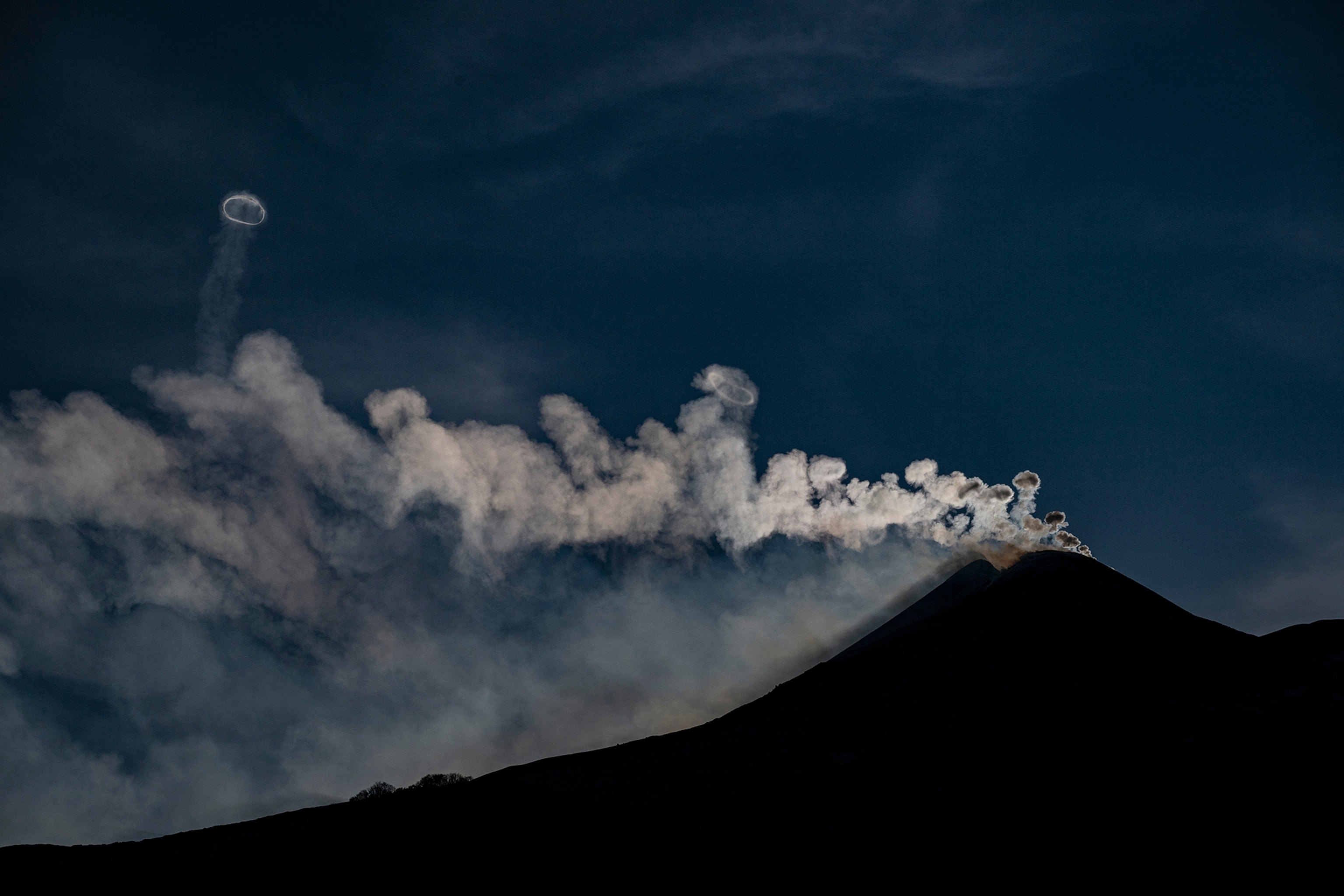 Mt Etna is silhouetted a stream of smoke veering to the right of the slope, you can see several smoke circles rising from the smoke one floats further than the others.