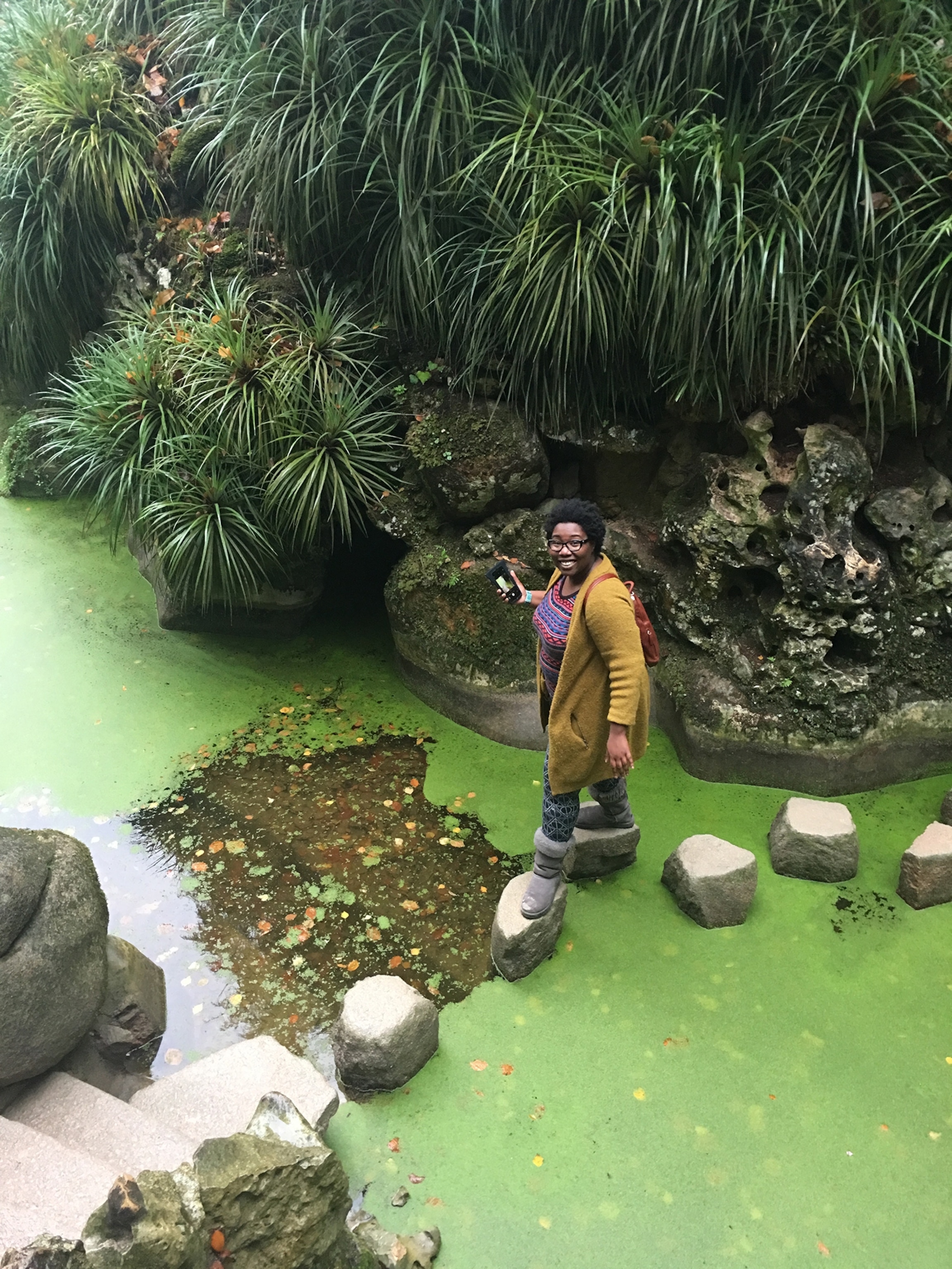 A woman woman with dark skin and an afro with glasses and a long yellow cardigan walks across stones of a green pond.