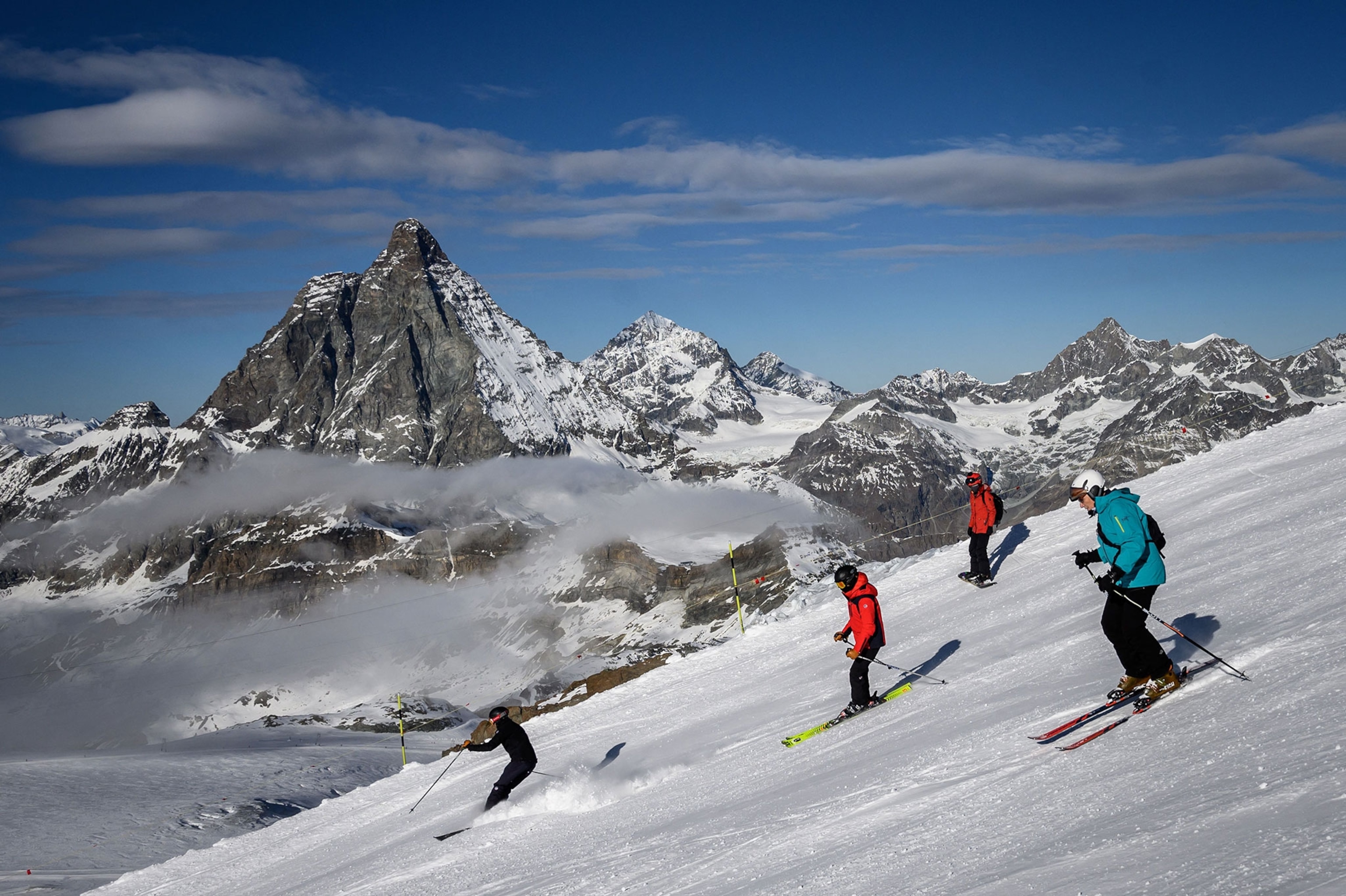 Four skiers, ski down a steep, snow-covered mountain side with the towering Matterhorn Mountain in the background.