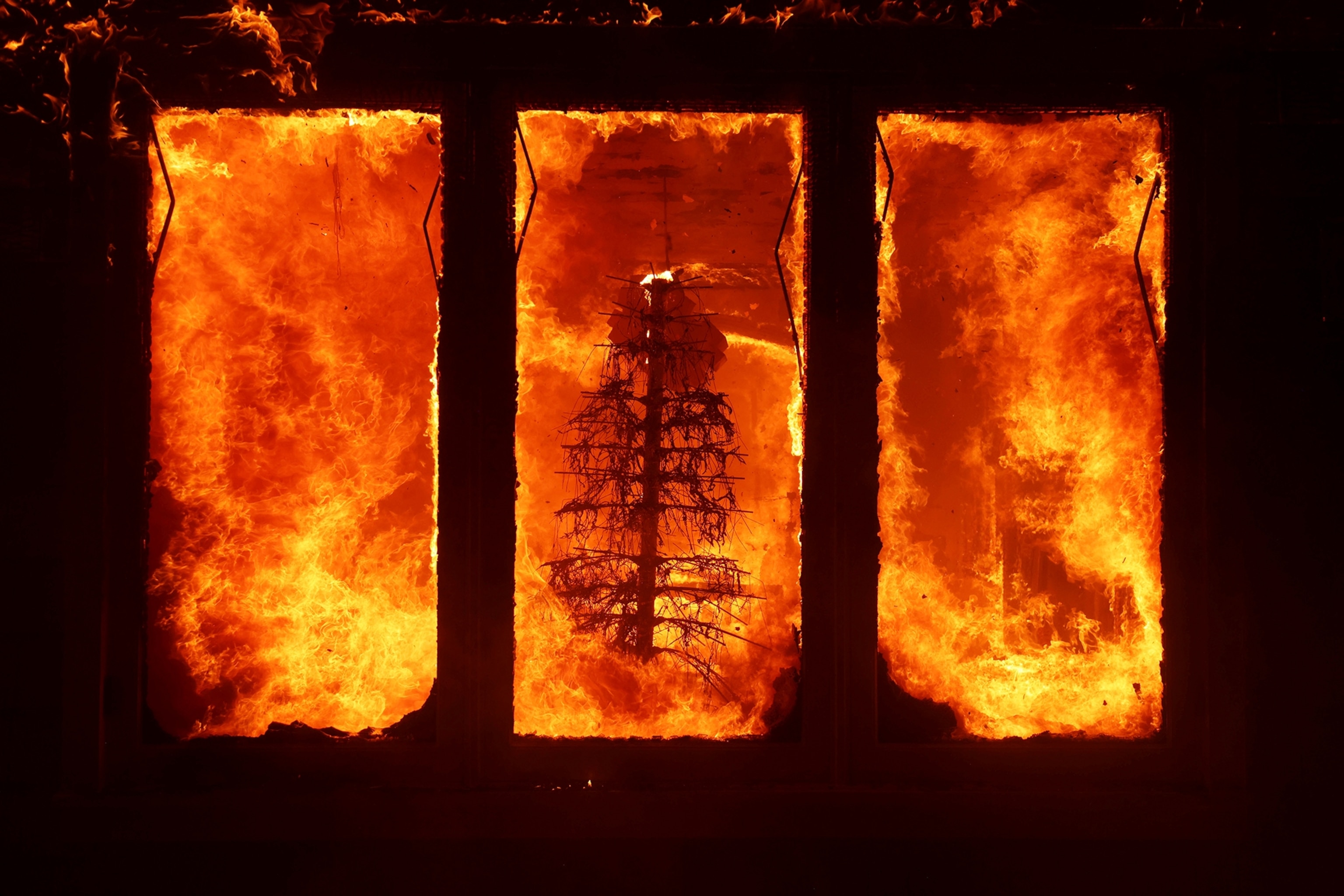 A Christmas tree is seen burning surrounded by flames behind three windows.