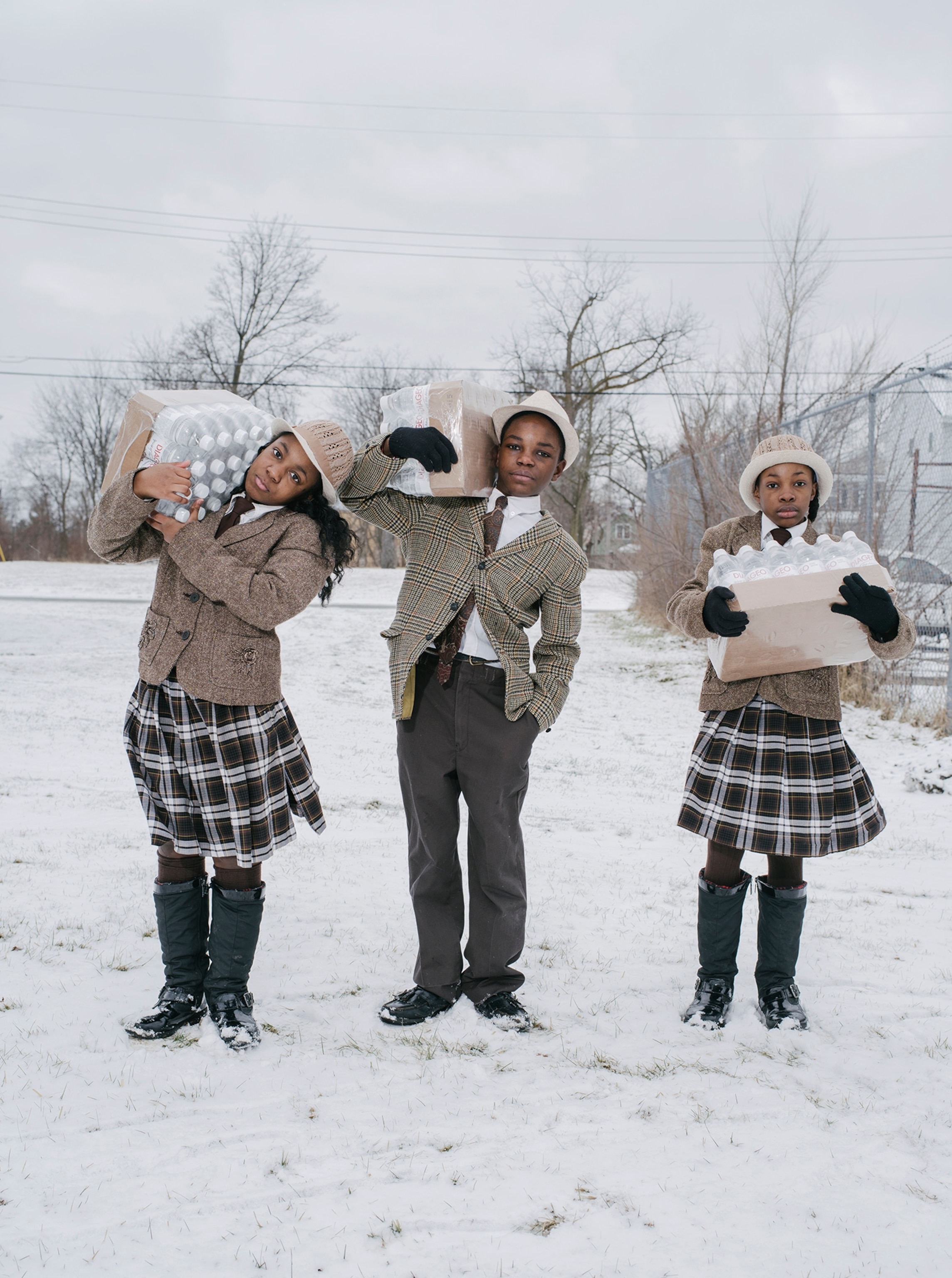 Three children in uniforms hold cases of water bottles outside in the snow