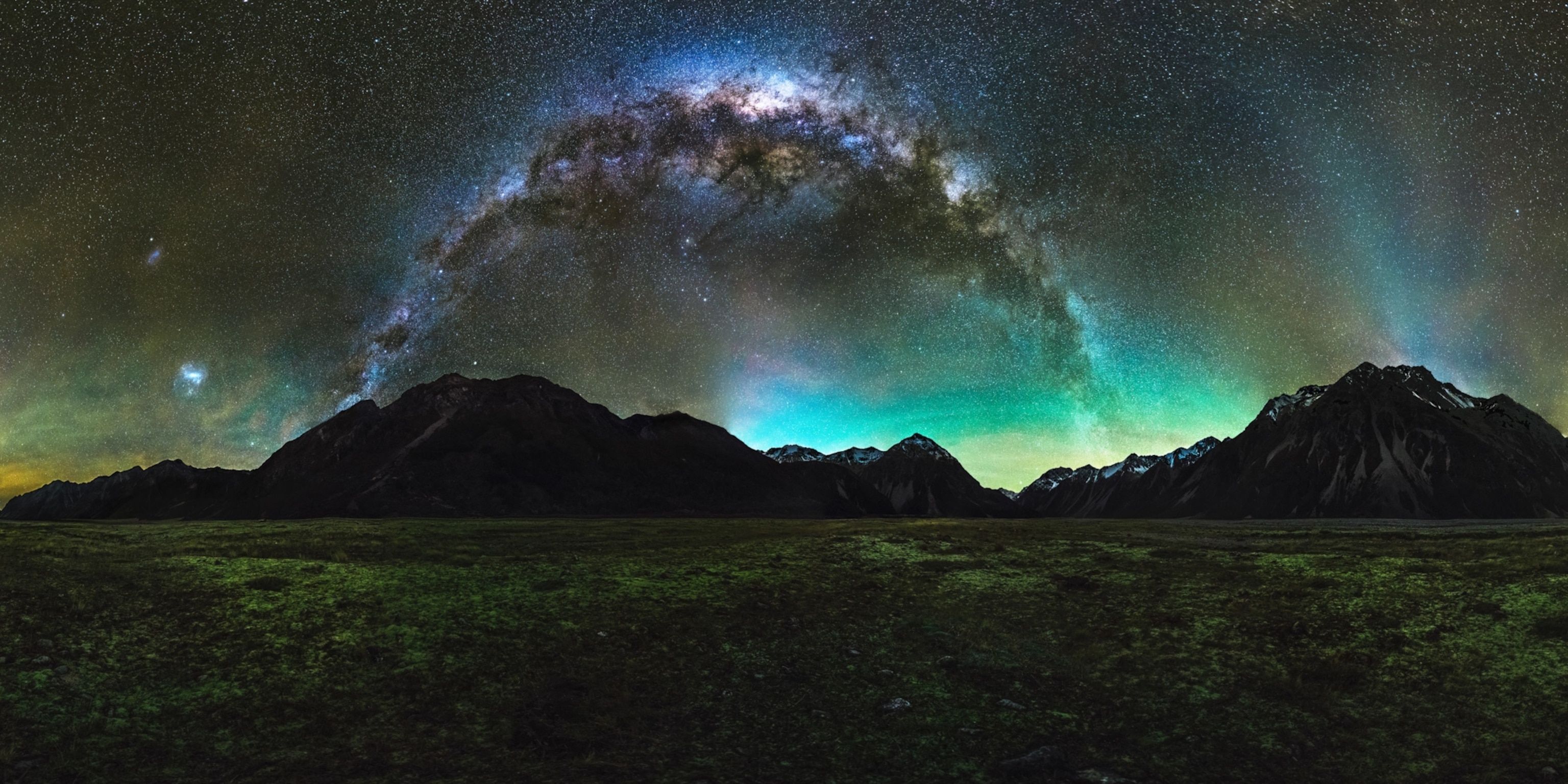 The milky way stretched across the sky before meeting with bands of green aurora light by mountains on the horizon.