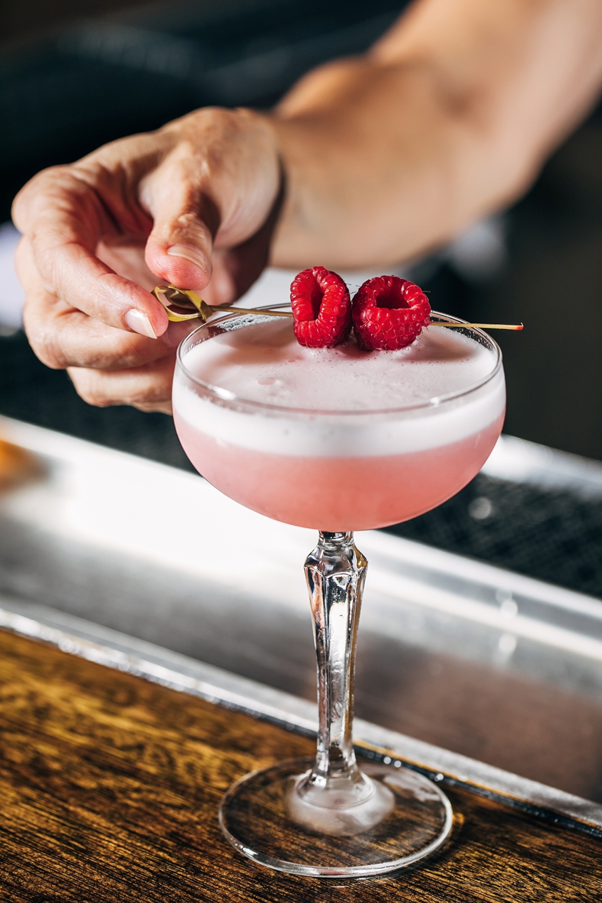 Close-up of a pink cocktail in a champagne coup with a male hand placing a cocktail pick with two raspberries onto the glass' rim.