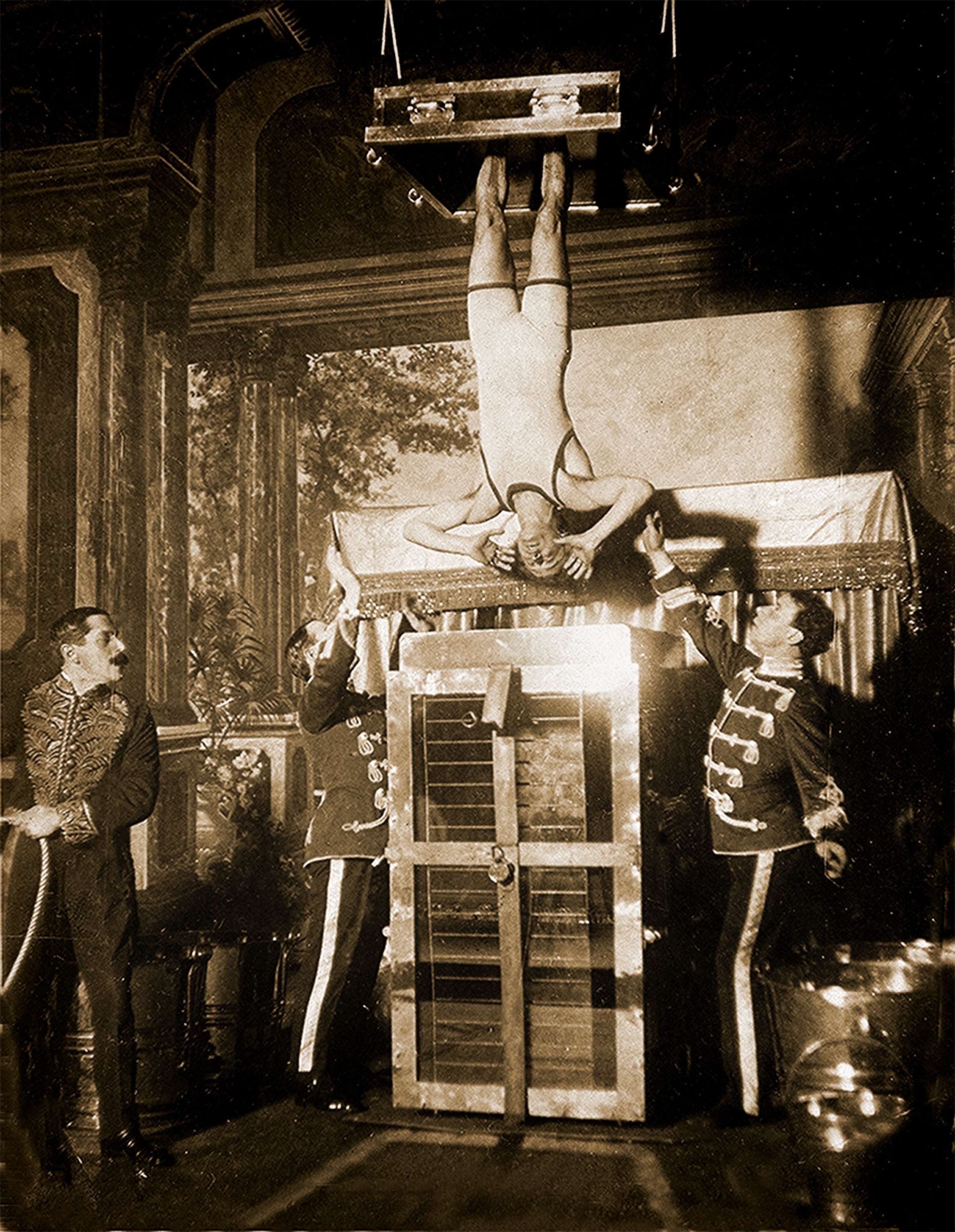 A photo shows Harry Houdini being lowered upside down into his water torture cell with 3 men's help