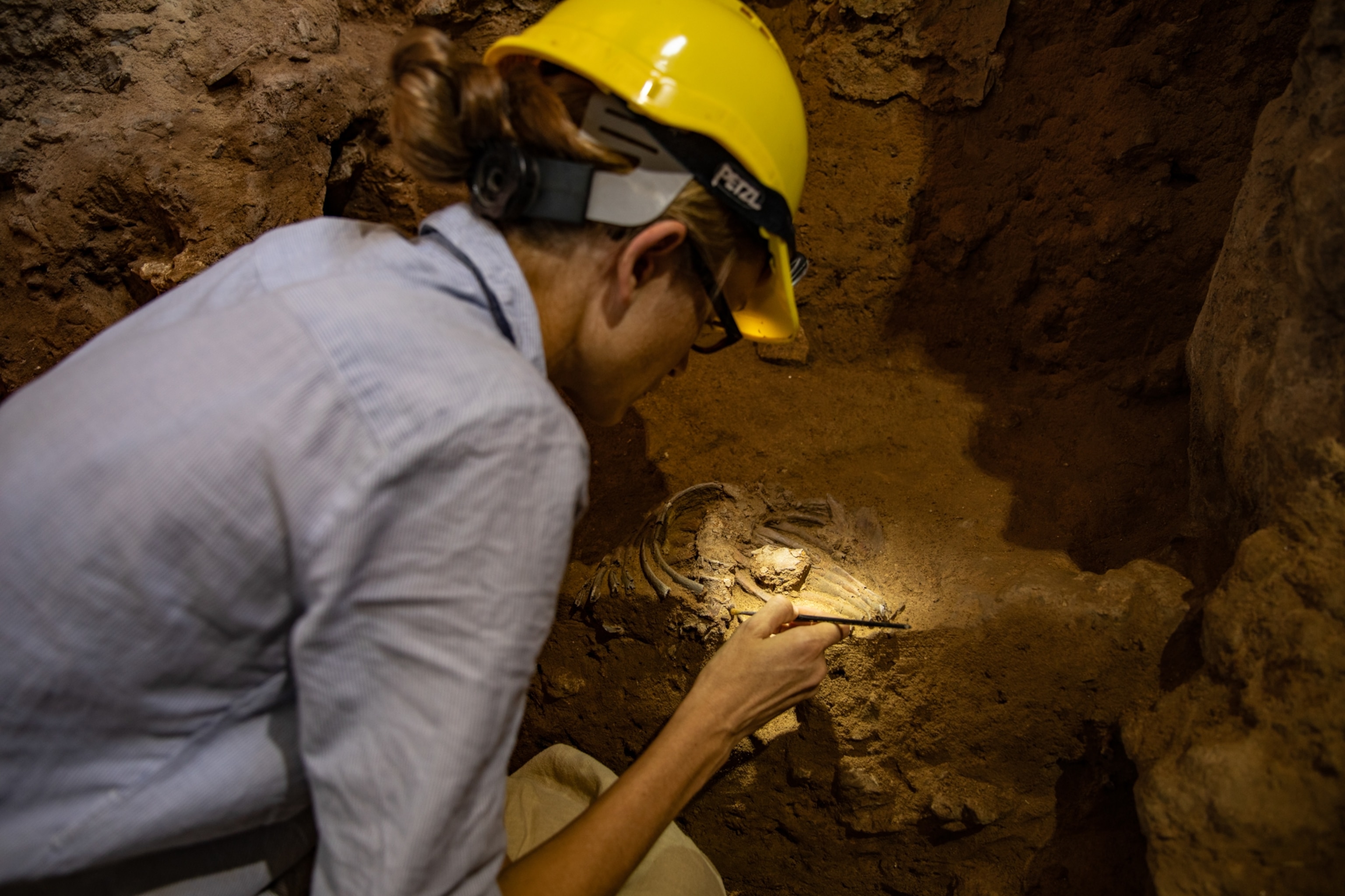 An archaeologist wearing a yellow hardhat, which shines a light down on the skeletal remains she is working to recover from the rocky cave floor.