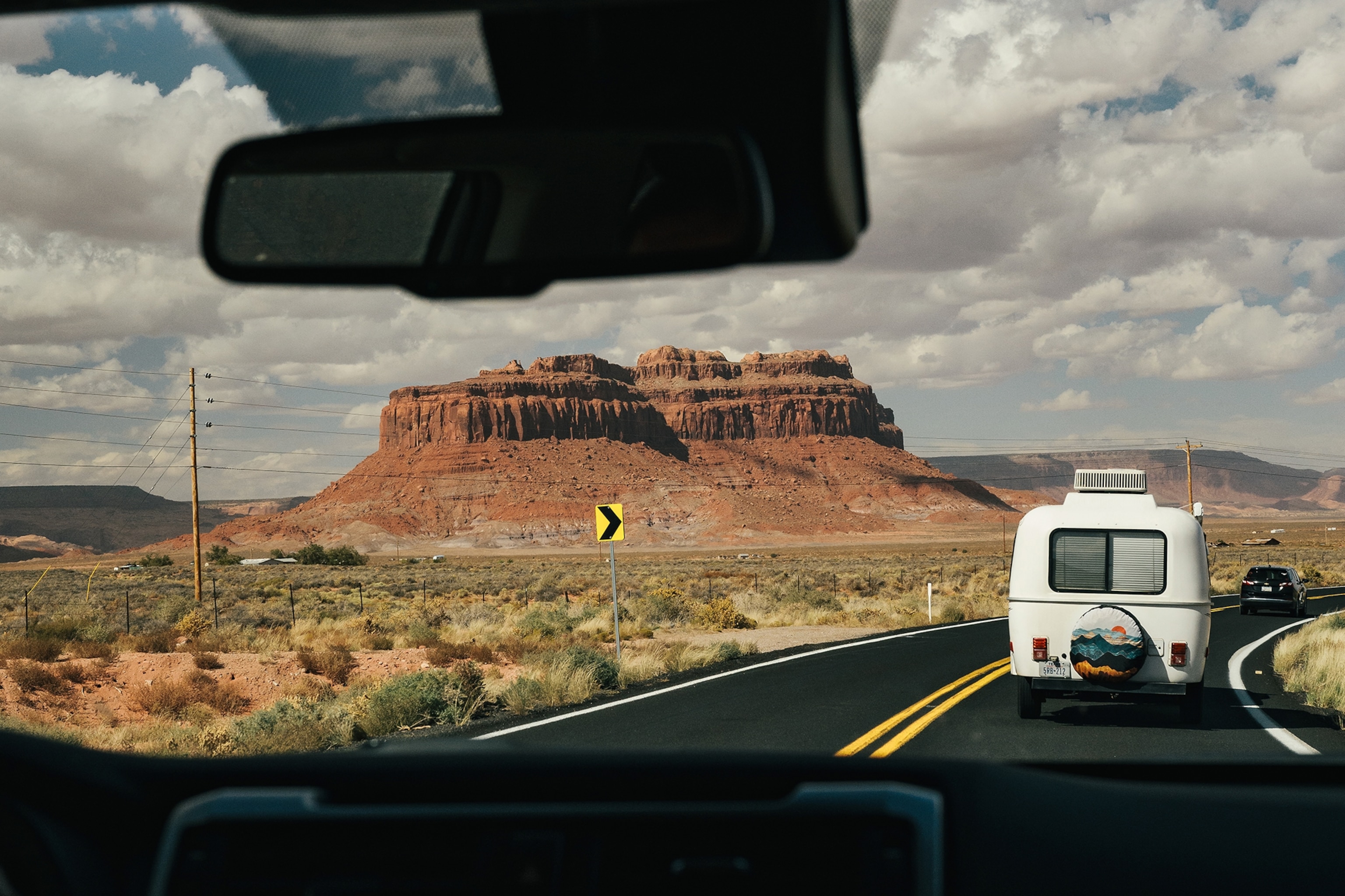 The view from the front seat of a car onto a busy winding desert road past Monument Valley, a national park of monolithic natural rock formations.